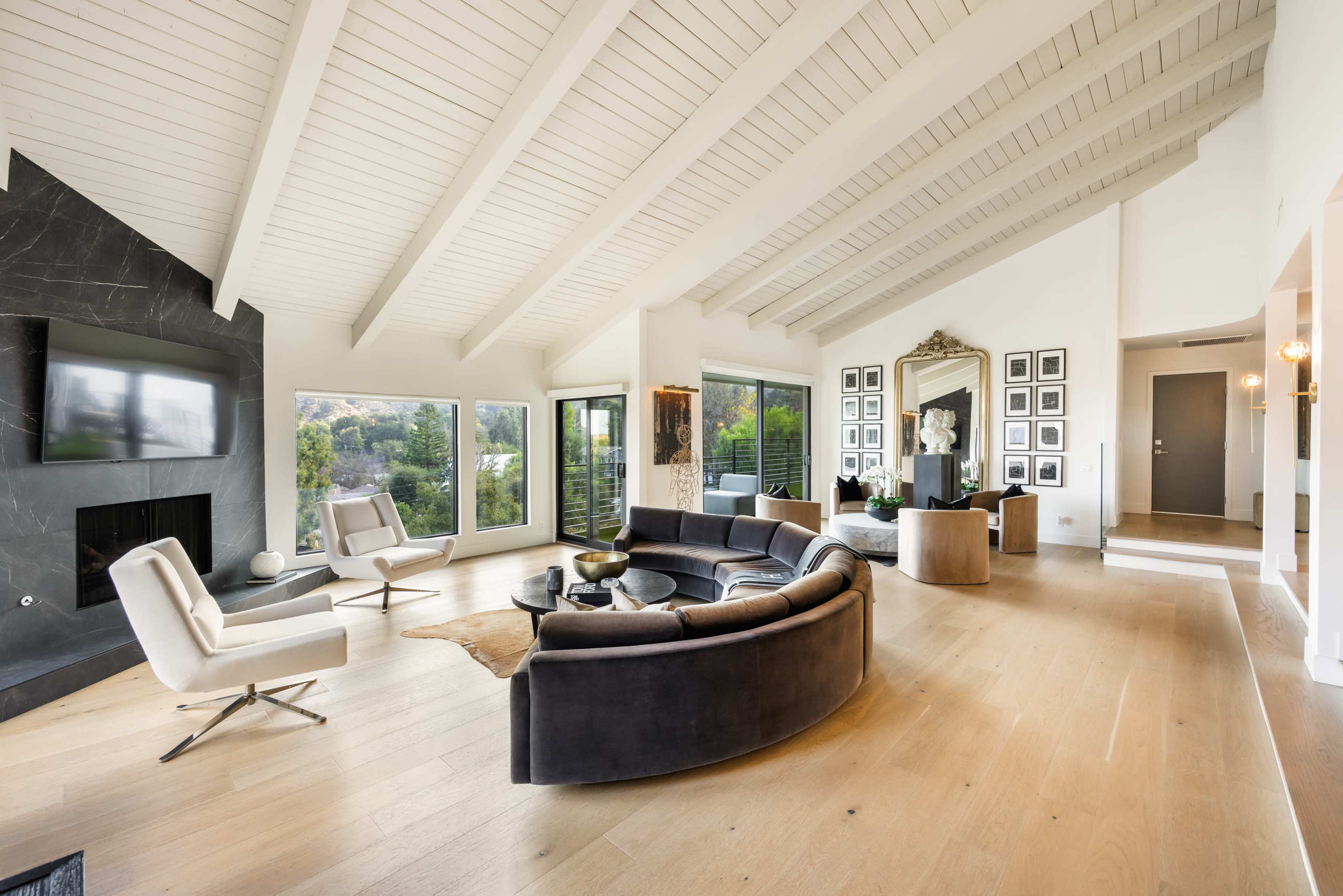 The spacious living room features a curved sectional sofa, two armchairs, and large windows overlooking greenery, all under a slanted ceiling with exposed beams.