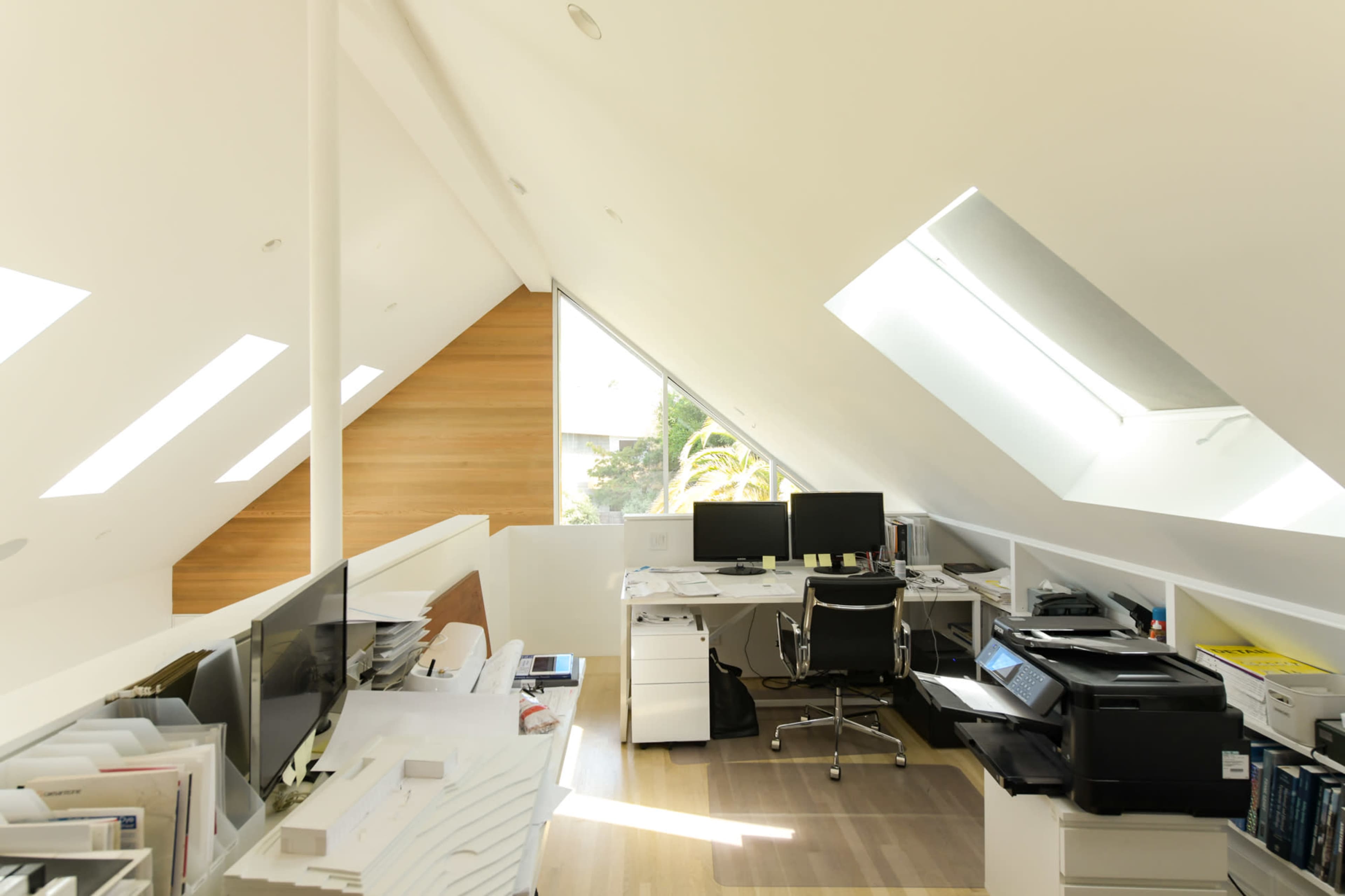 A modern attic office space featuring two computer workstations, a printer, and large skylights that illuminate the area.