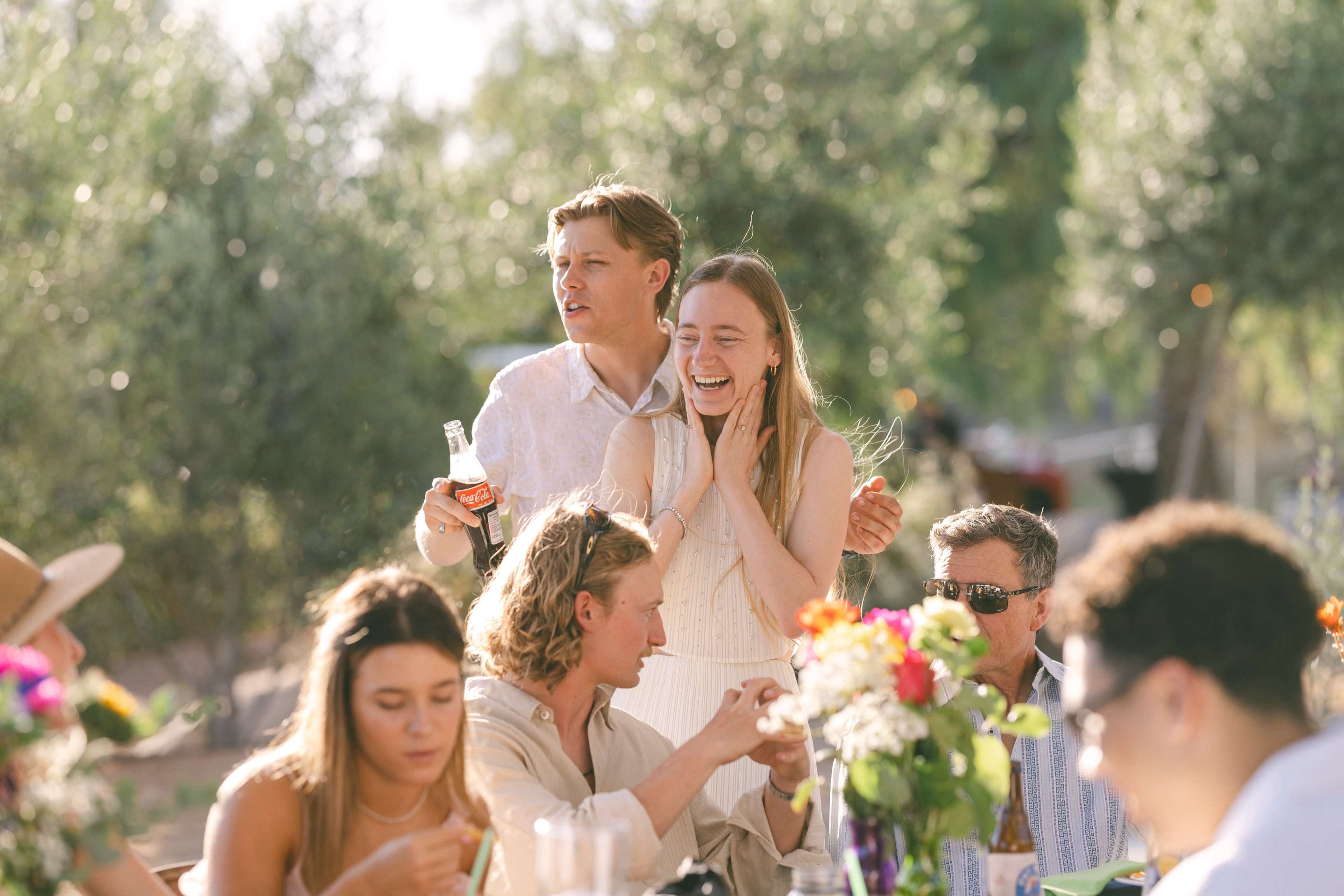 A group of people enjoys a lively outdoor gathering, with several individuals laughing and interacting at a table adorned with flowers.