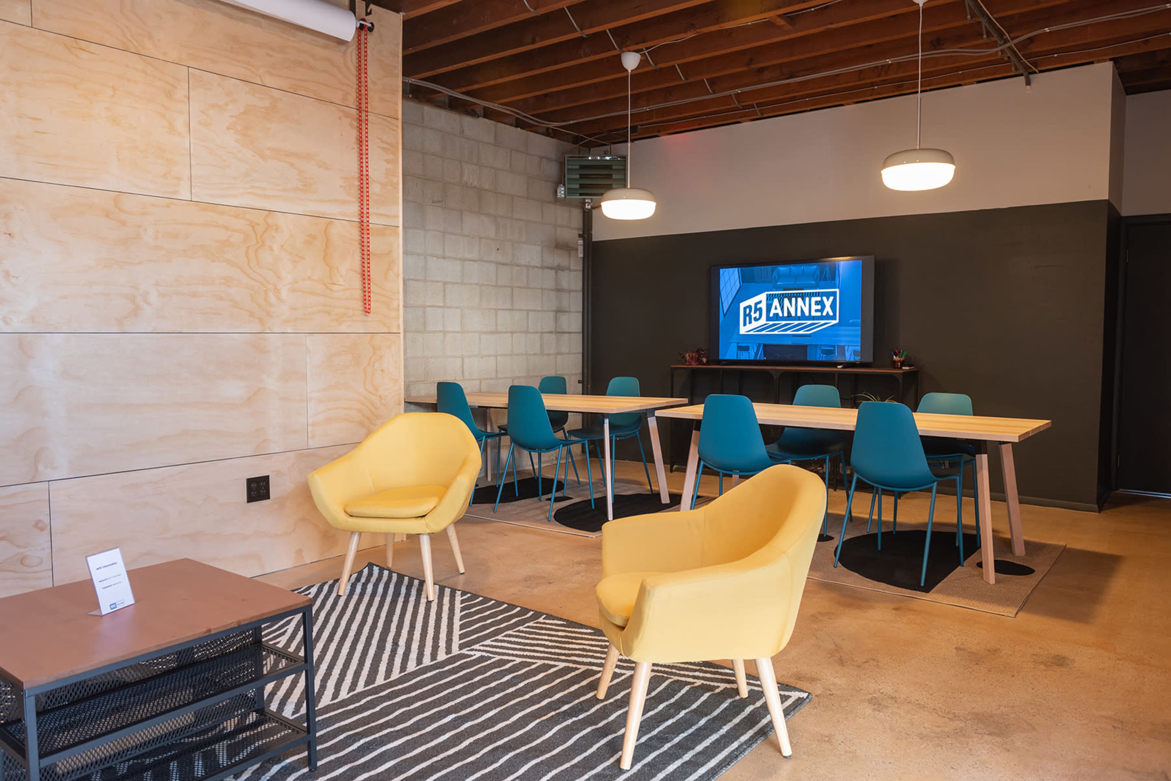 A modern meeting room features a long table surrounded by teal chairs, two yellow armchairs, and a television displaying the "R5 Annex" logo.