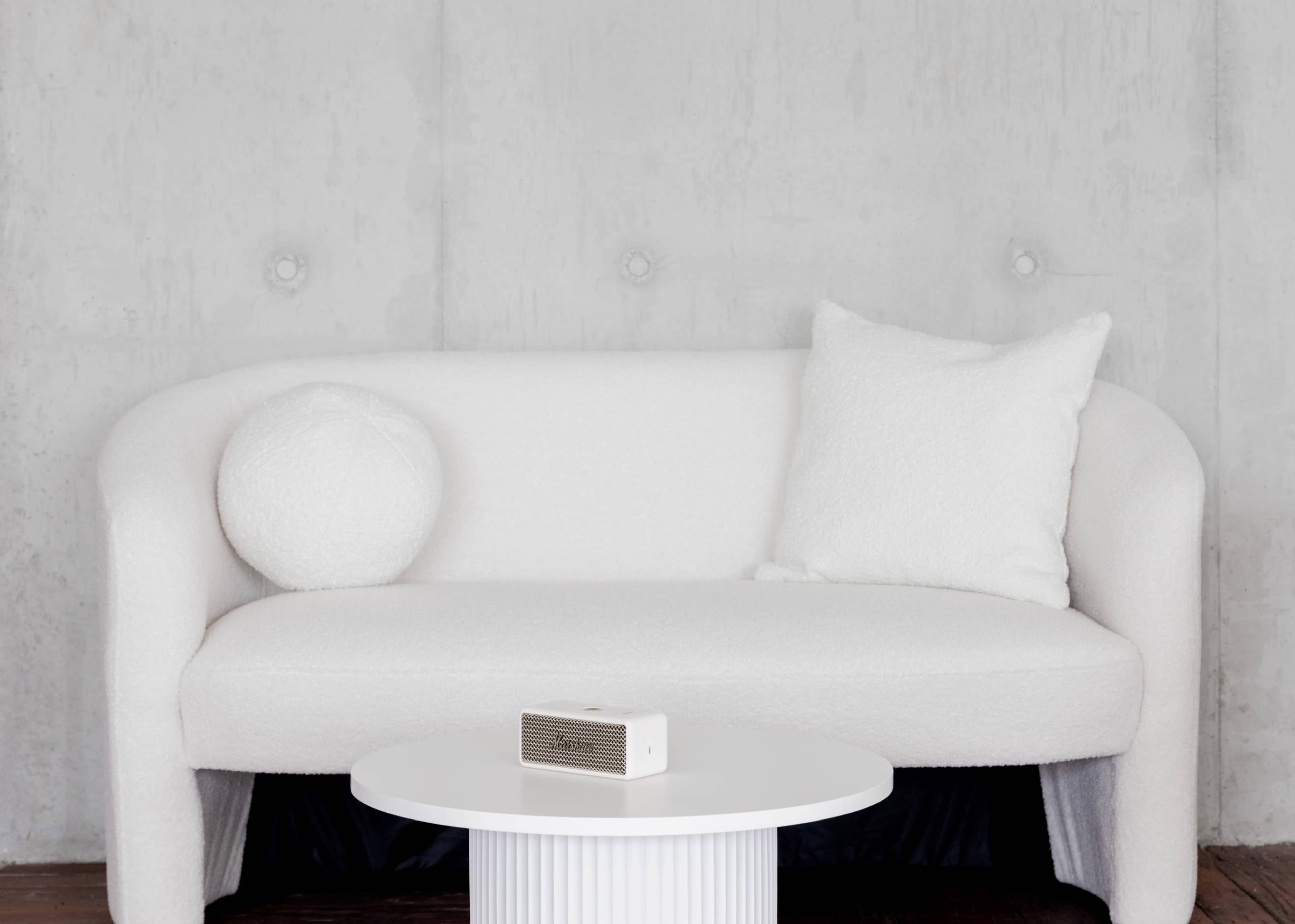 A white, fluffy sofa and a round cushion sit beside a white table with a small speaker on it against a gray wall.