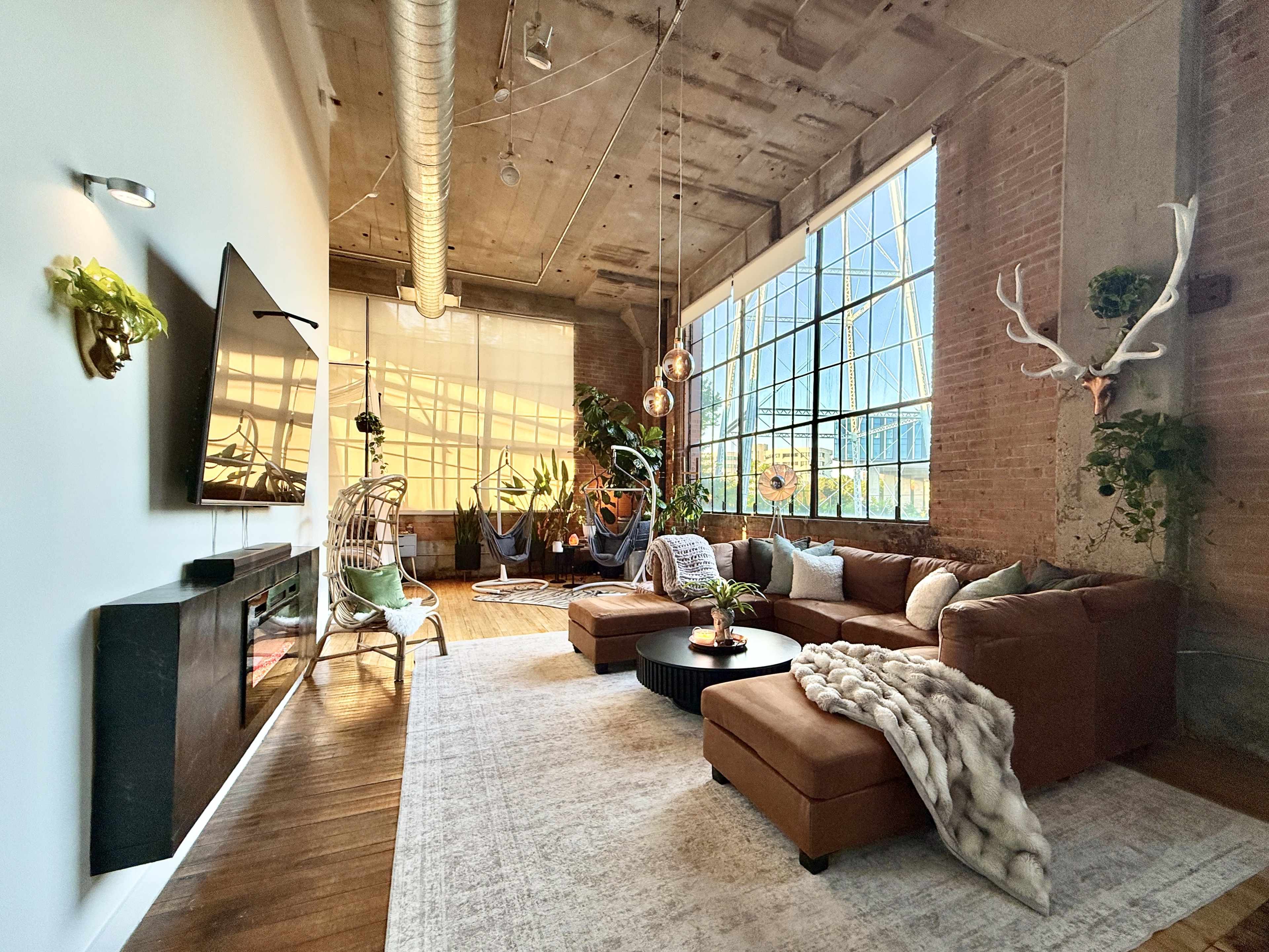 The image shows a spacious living room with large windows, exposed brick walls, a sectional sofa, and various indoor plants.