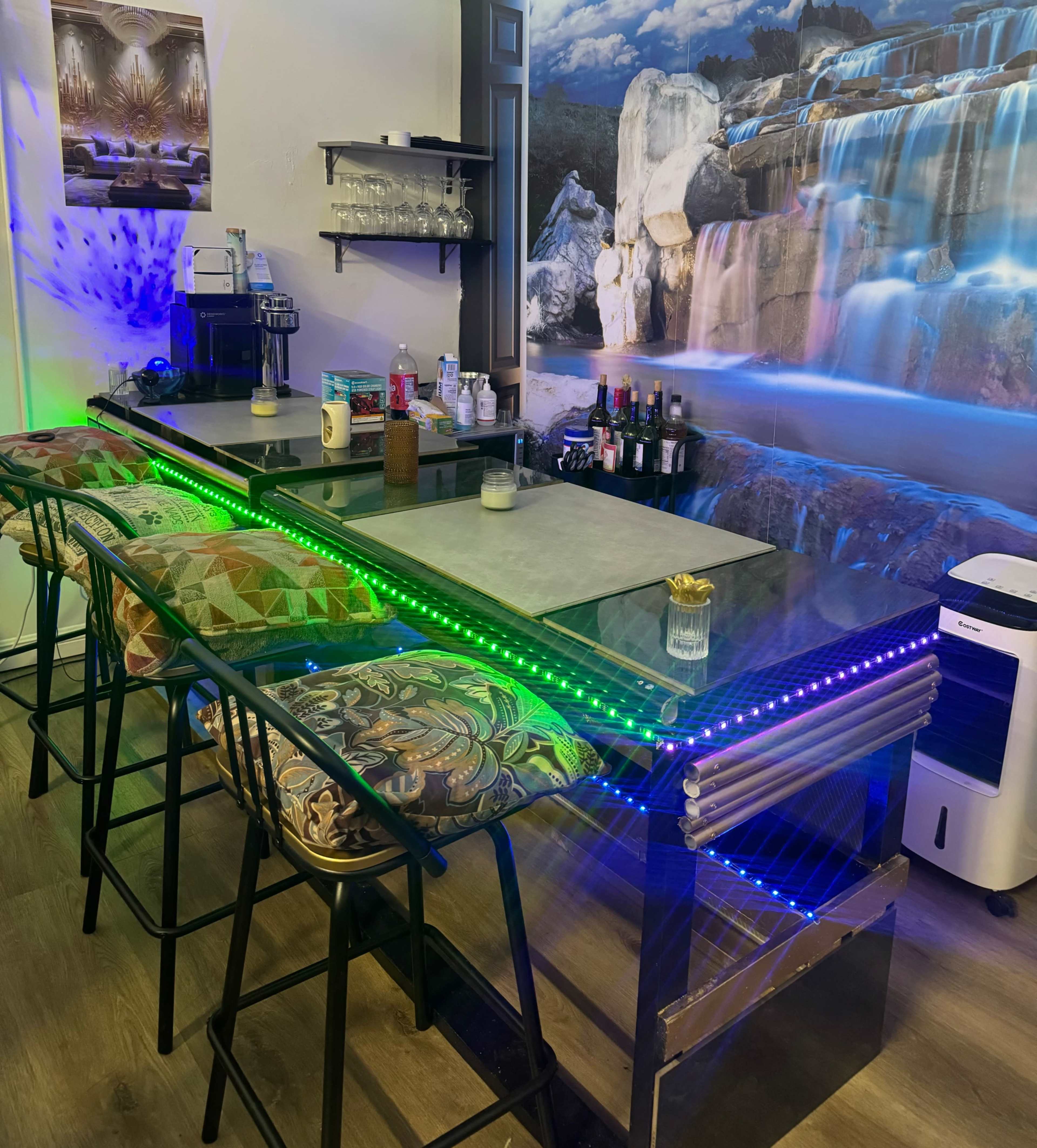 The image shows a modern bar area with LED lighting, a counter, stools with patterned upholstery, and a backdrop of a waterfall mural.