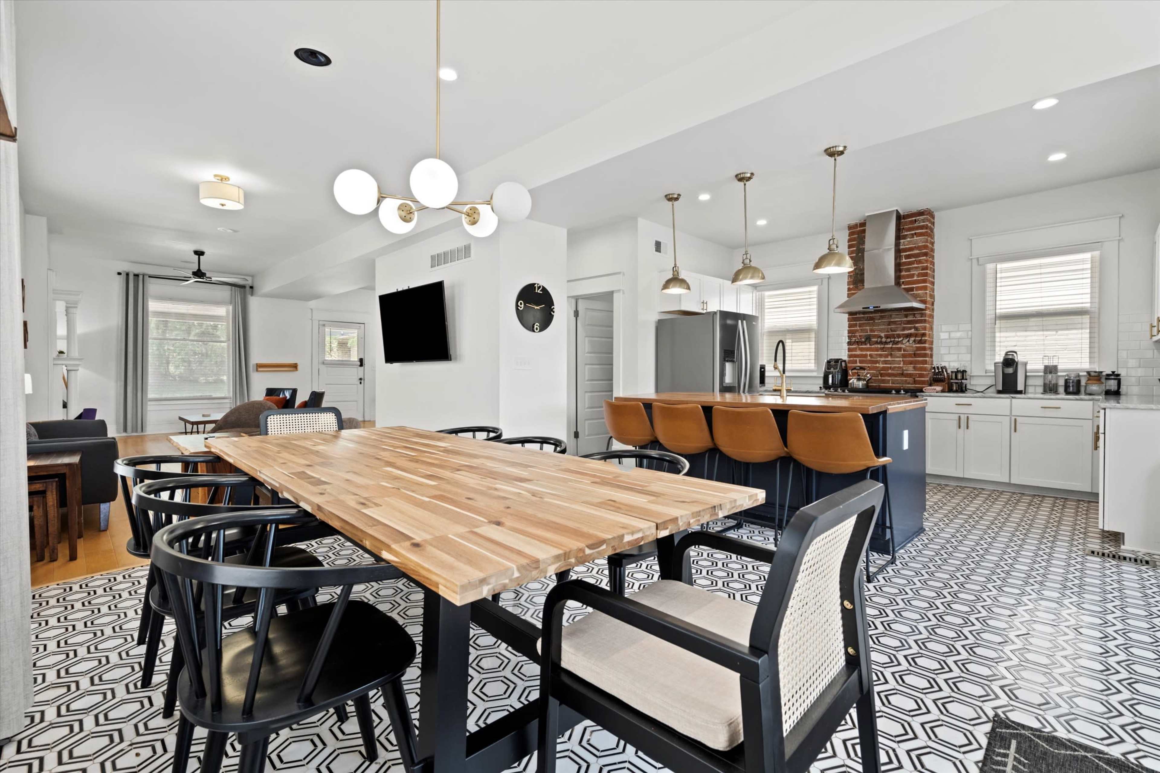 The image shows a modern kitchen and dining area featuring a wooden dining table, black chairs, and a kitchen with stainless steel appliances and a patterned tile floor.