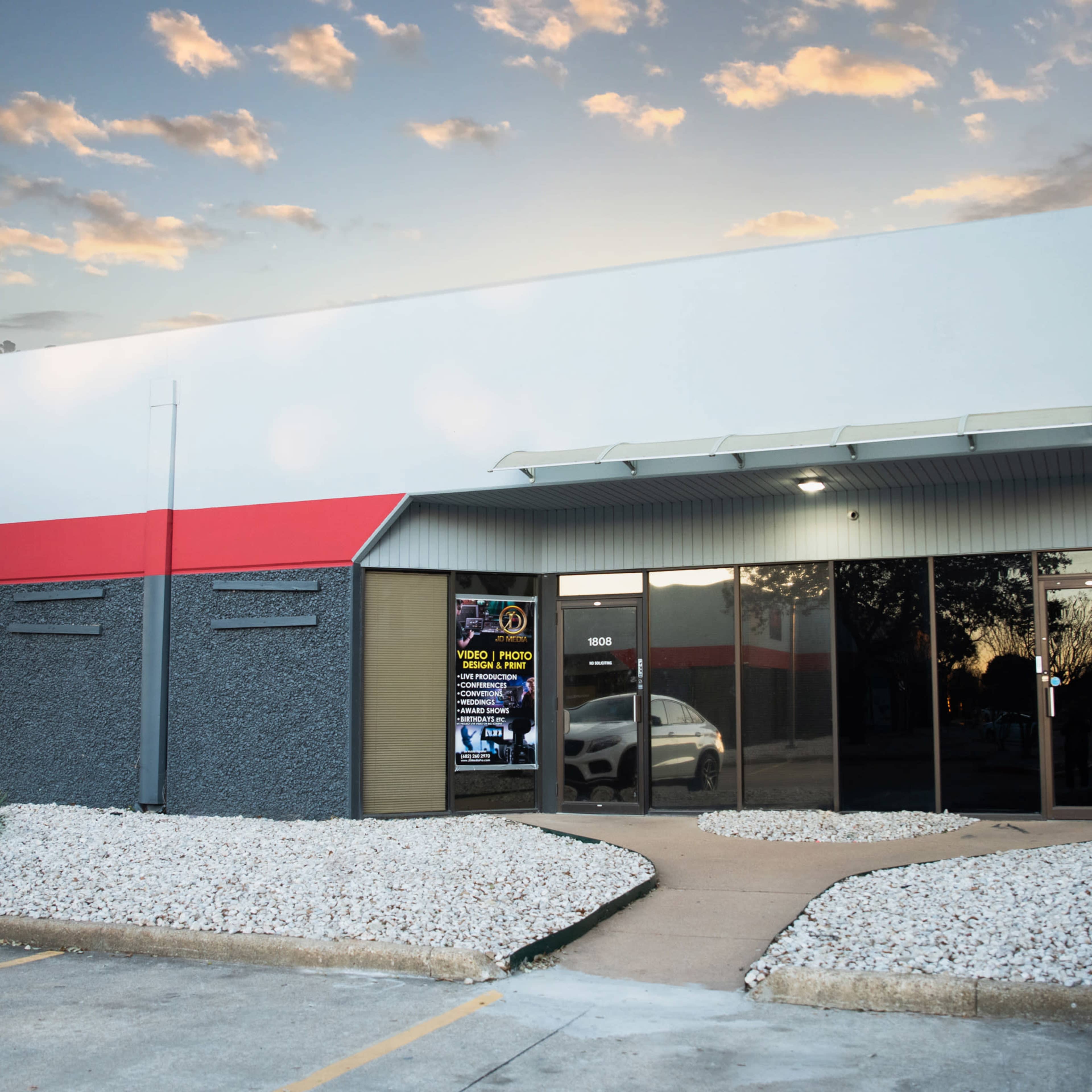 The image shows a commercial building with a red and gray exterior, featuring a front entrance and white gravel landscaping.