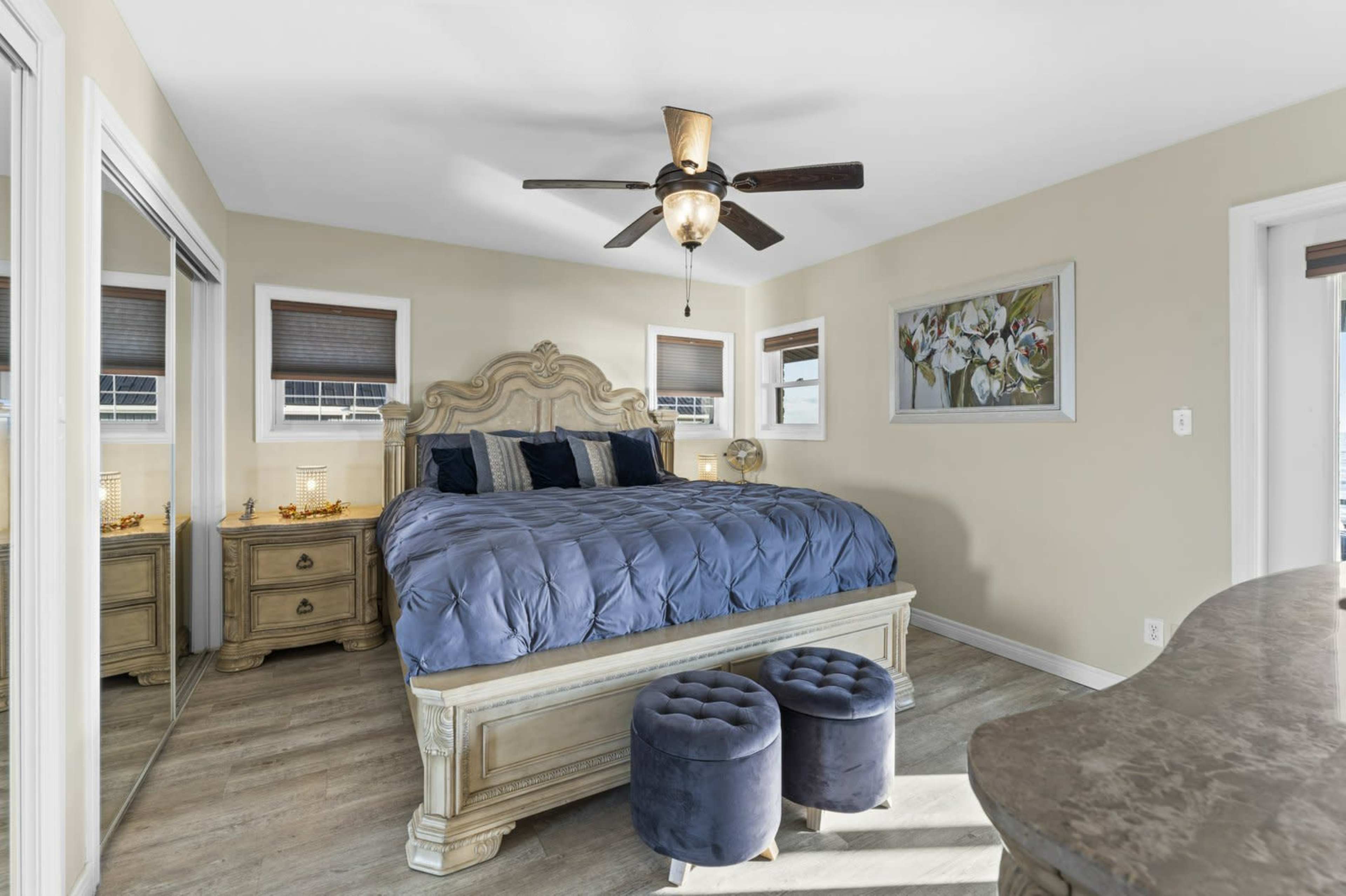 The image shows a well-furnished bedroom featuring a large bed with a tufted blue comforter, a decorative headboard, two upholstered ottomans, and a ceiling fan.