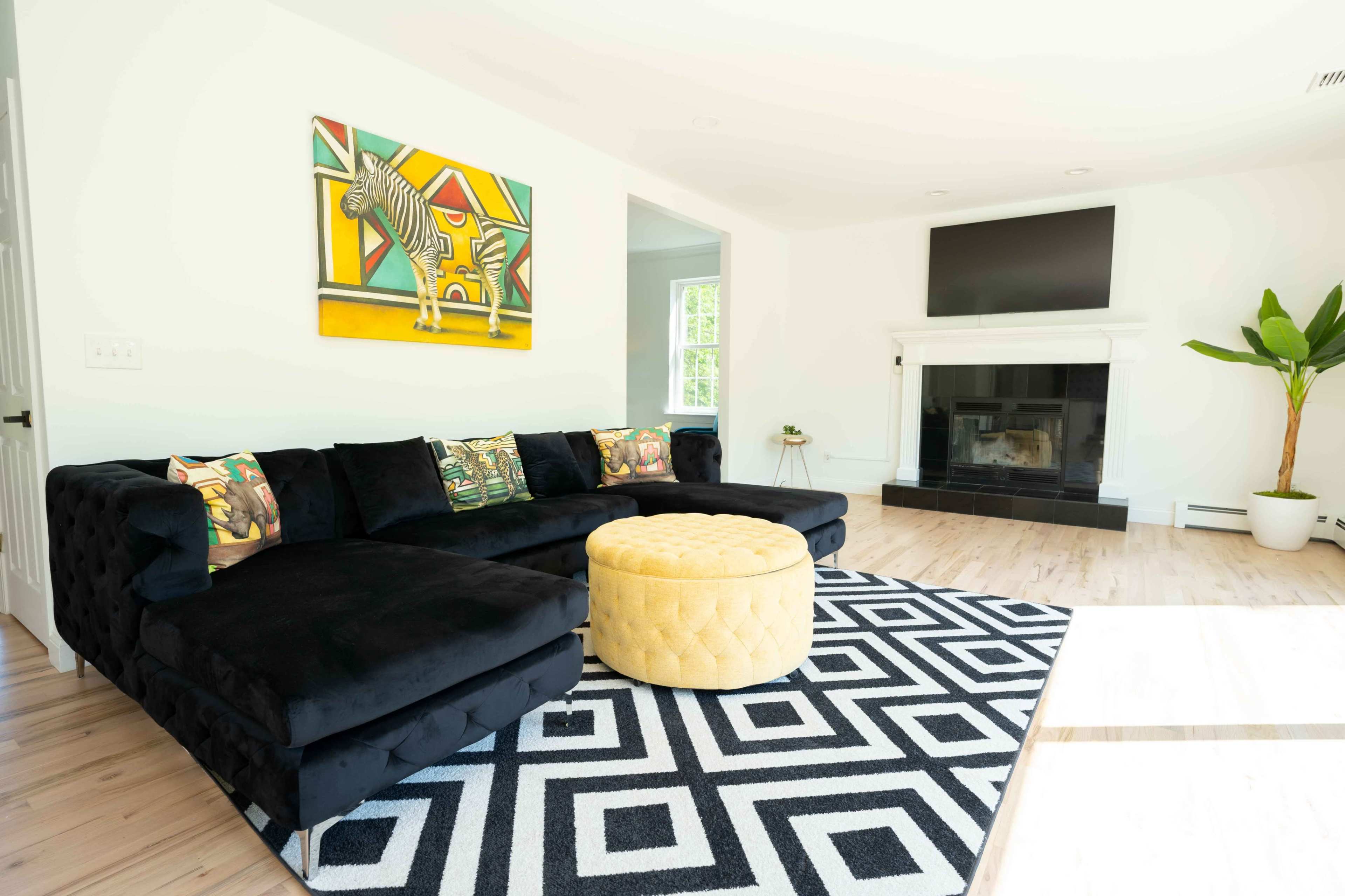 A modern living room features a black sectional sofa, a circular yellow ottoman, geometric black-and-white rug, a flat-screen TV mounted on the wall, and a vibrant artwork above the sofa.