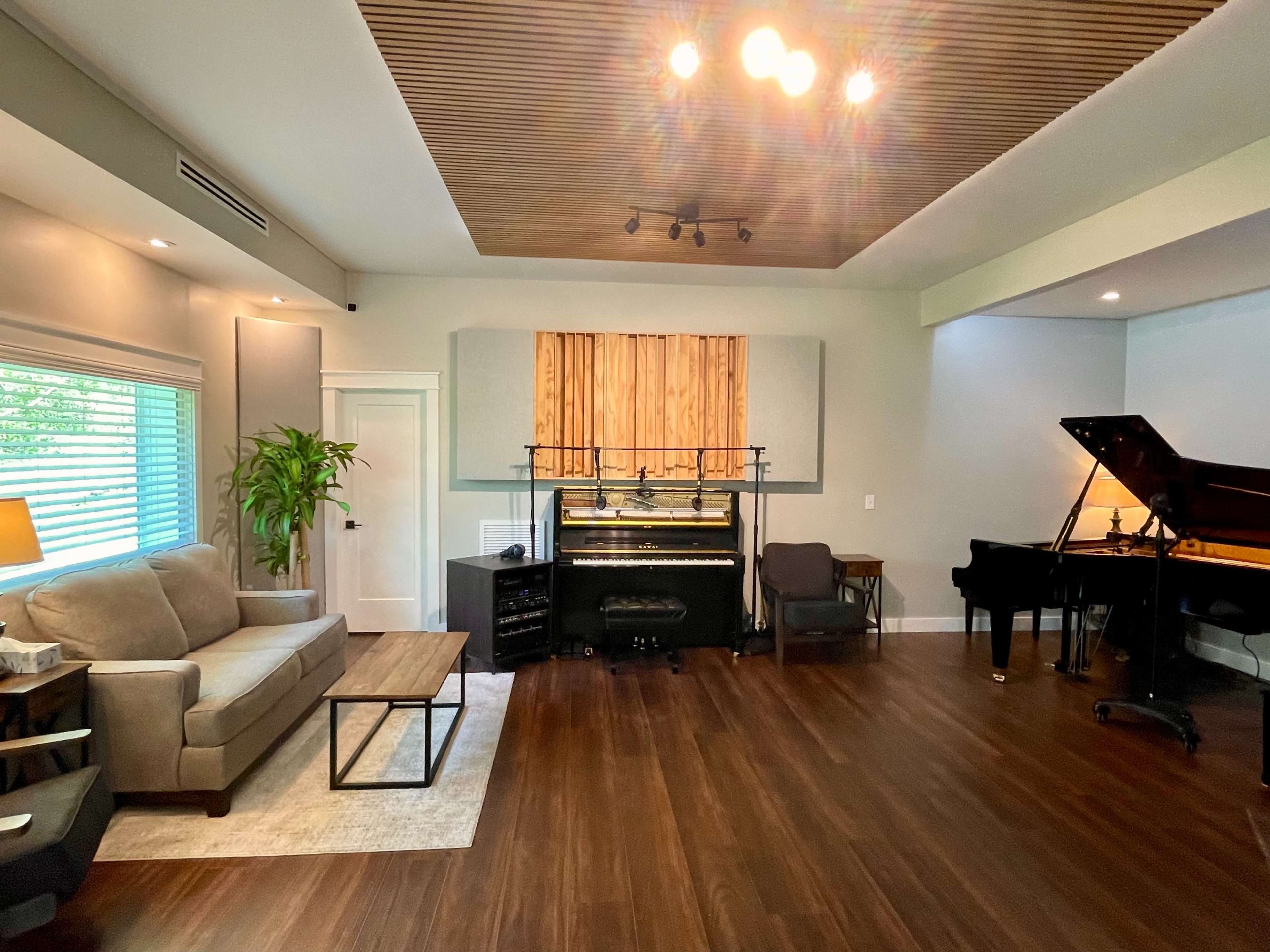 A spacious room features a piano, a couch, a coffee table, and large windows with natural light.