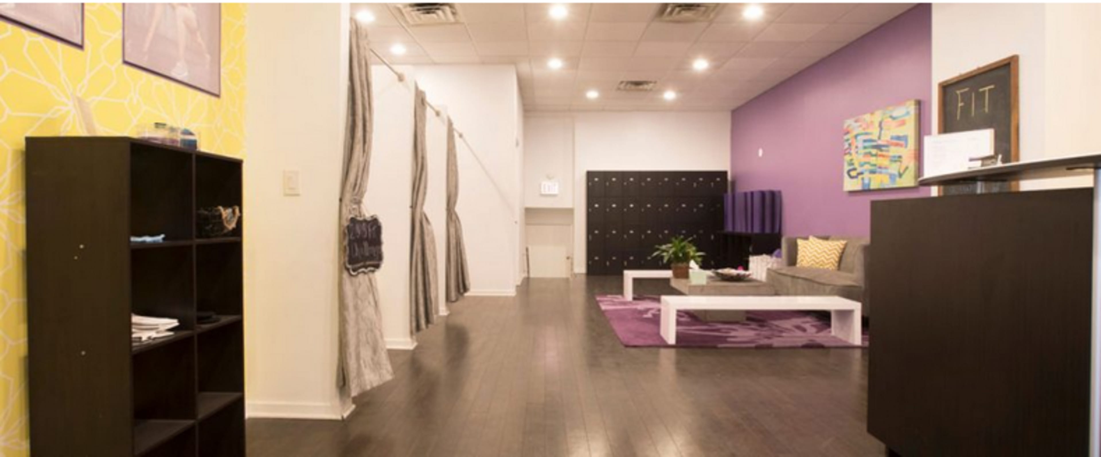 The image shows a modern interior space featuring a waiting area with a gray sofa, a purple accent wall, and a row of lockers in the background.