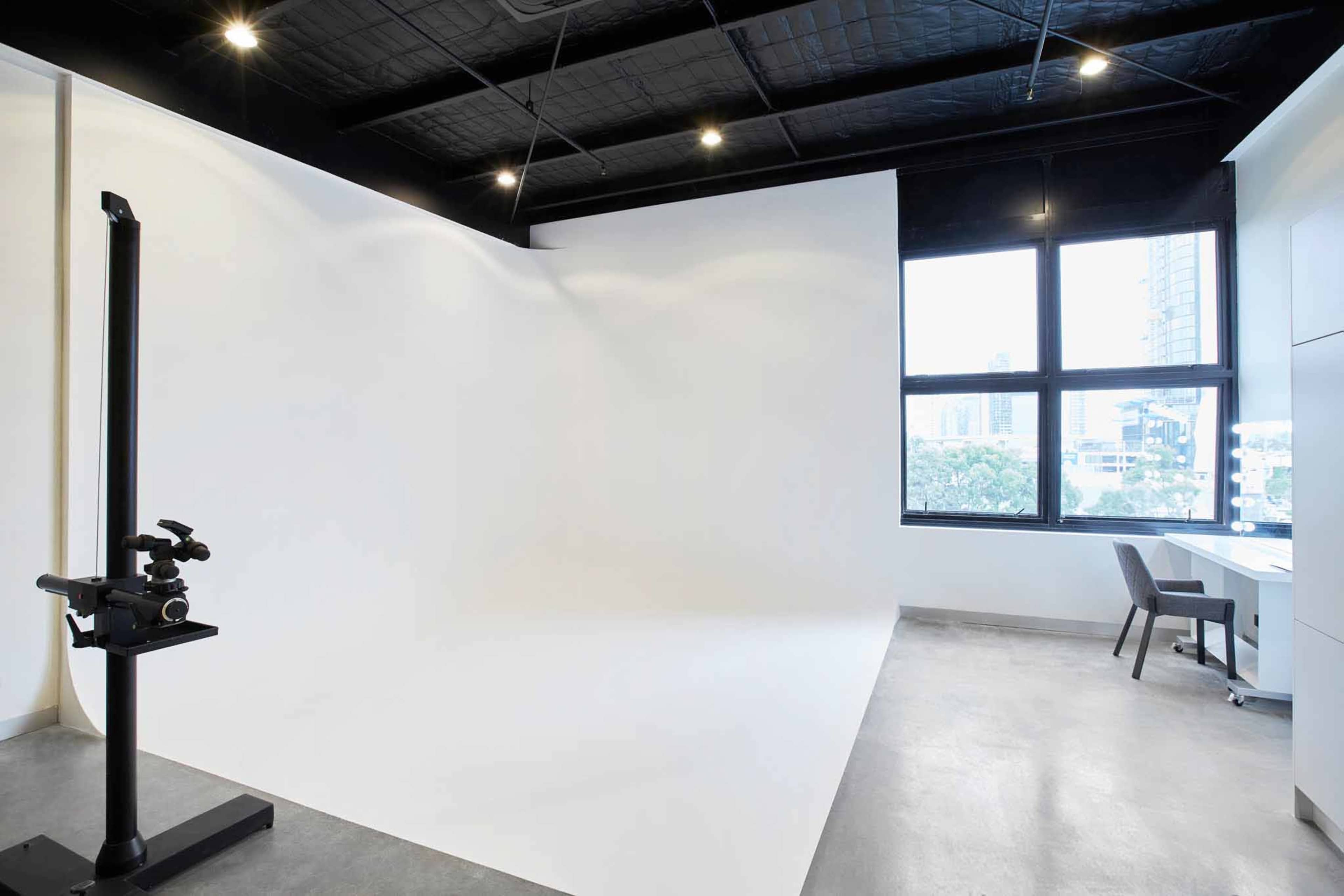 A spacious photography studio with a white backdrop, large windows, and a simple desk with a chair.