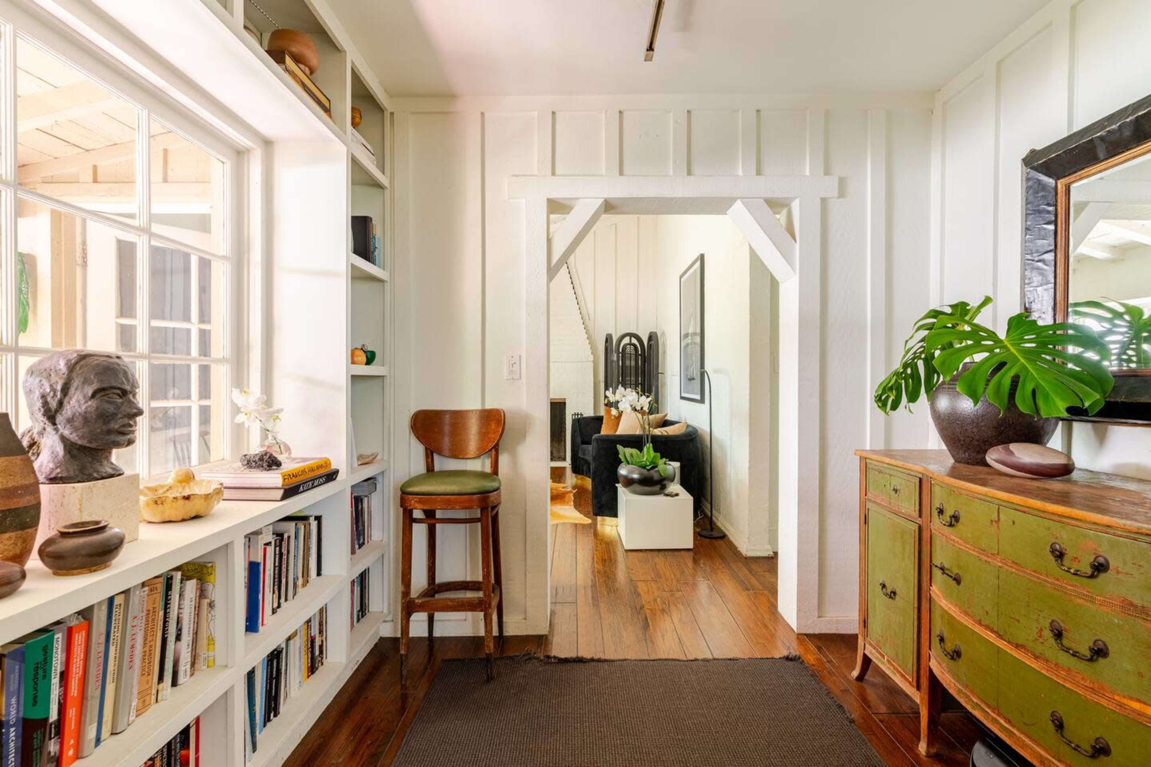 A well-lit hallway features bookshelves, a chair, and a decorative green cabinet leading to a living area with plants and artwork.
