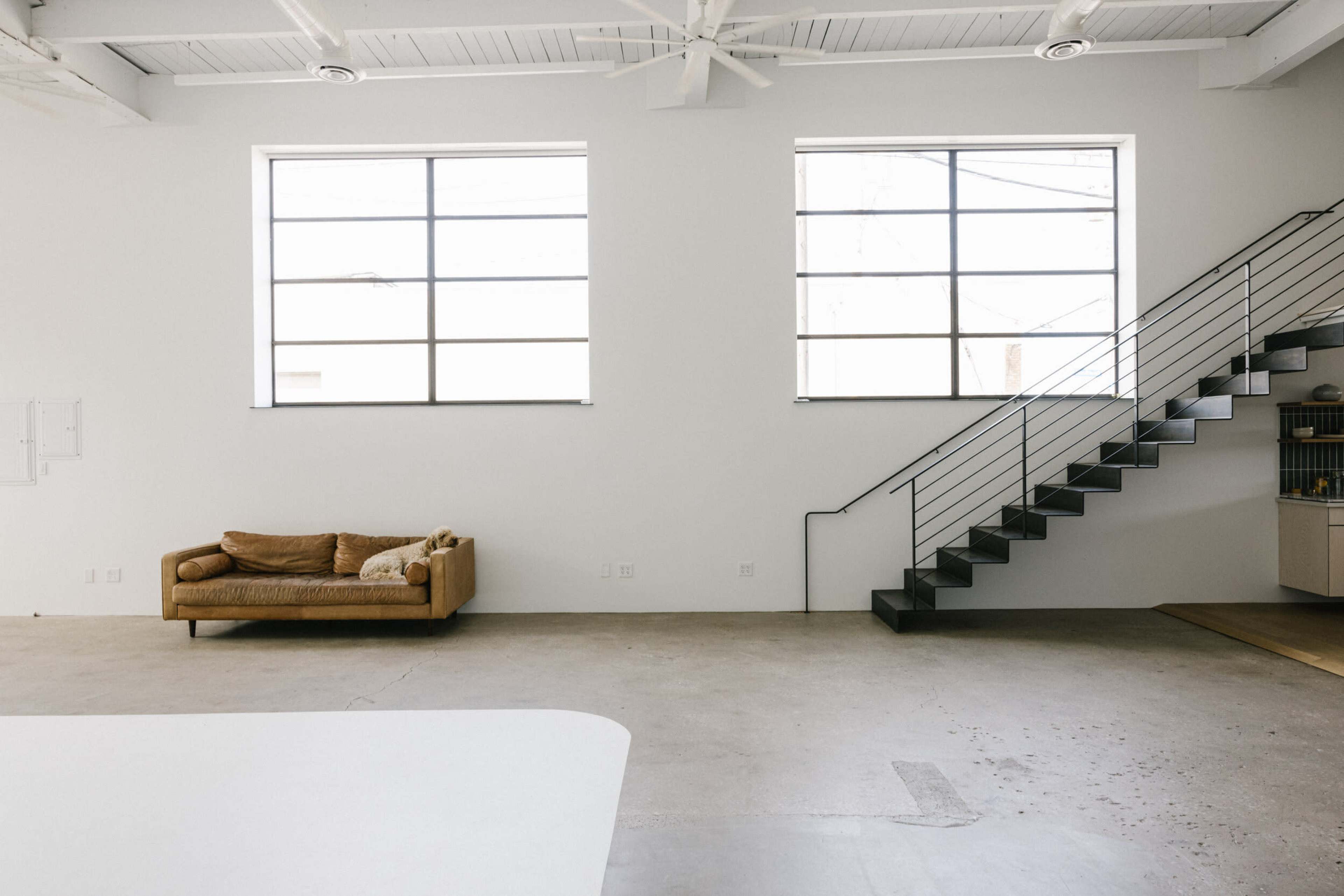 The scene features a minimalist interior with a brown couch against a wall and a staircase leading to an upper level.