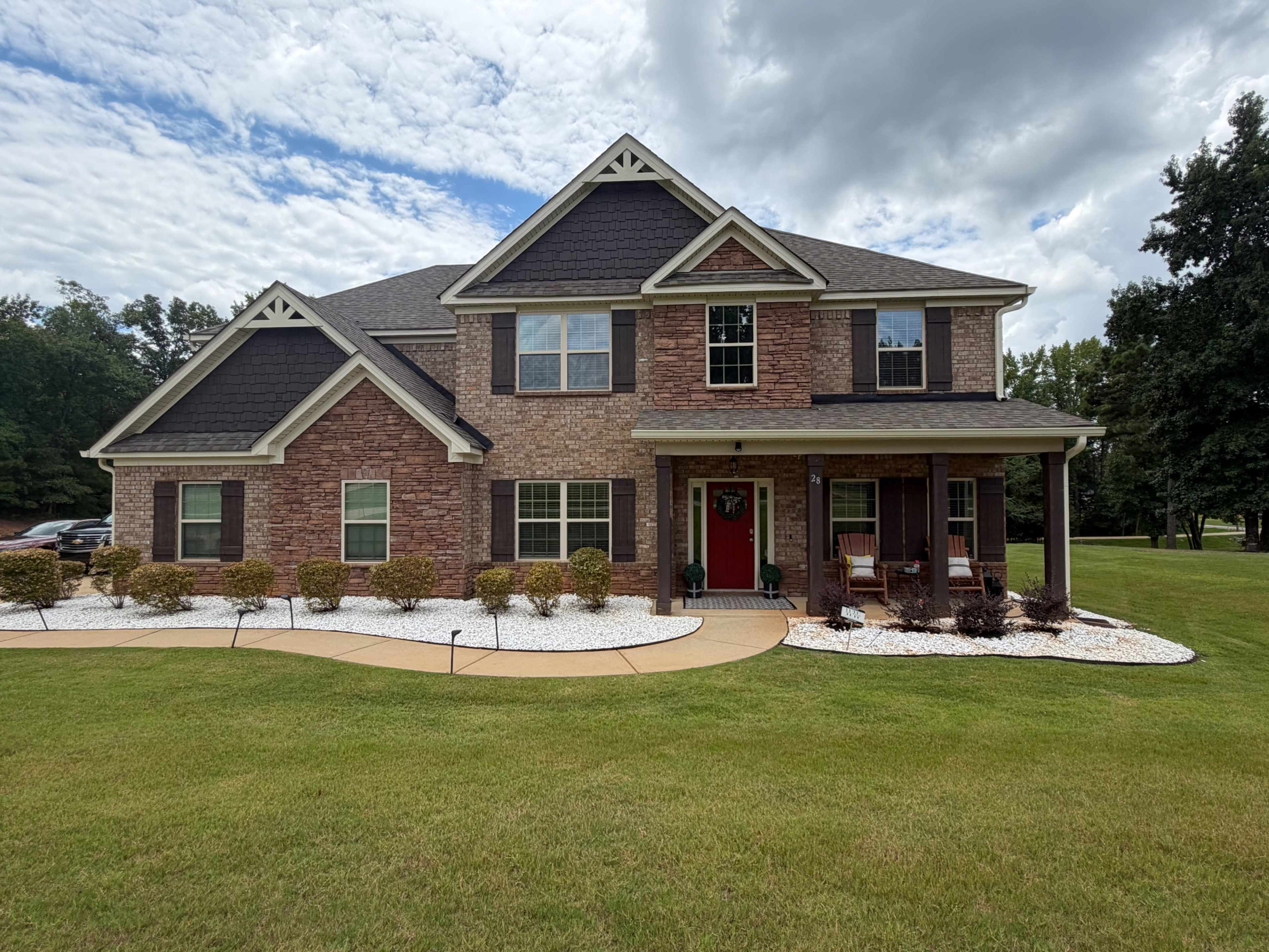 A large two-story brick house with a red front door and white stone landscaping is surrounded by a well-maintained lawn.