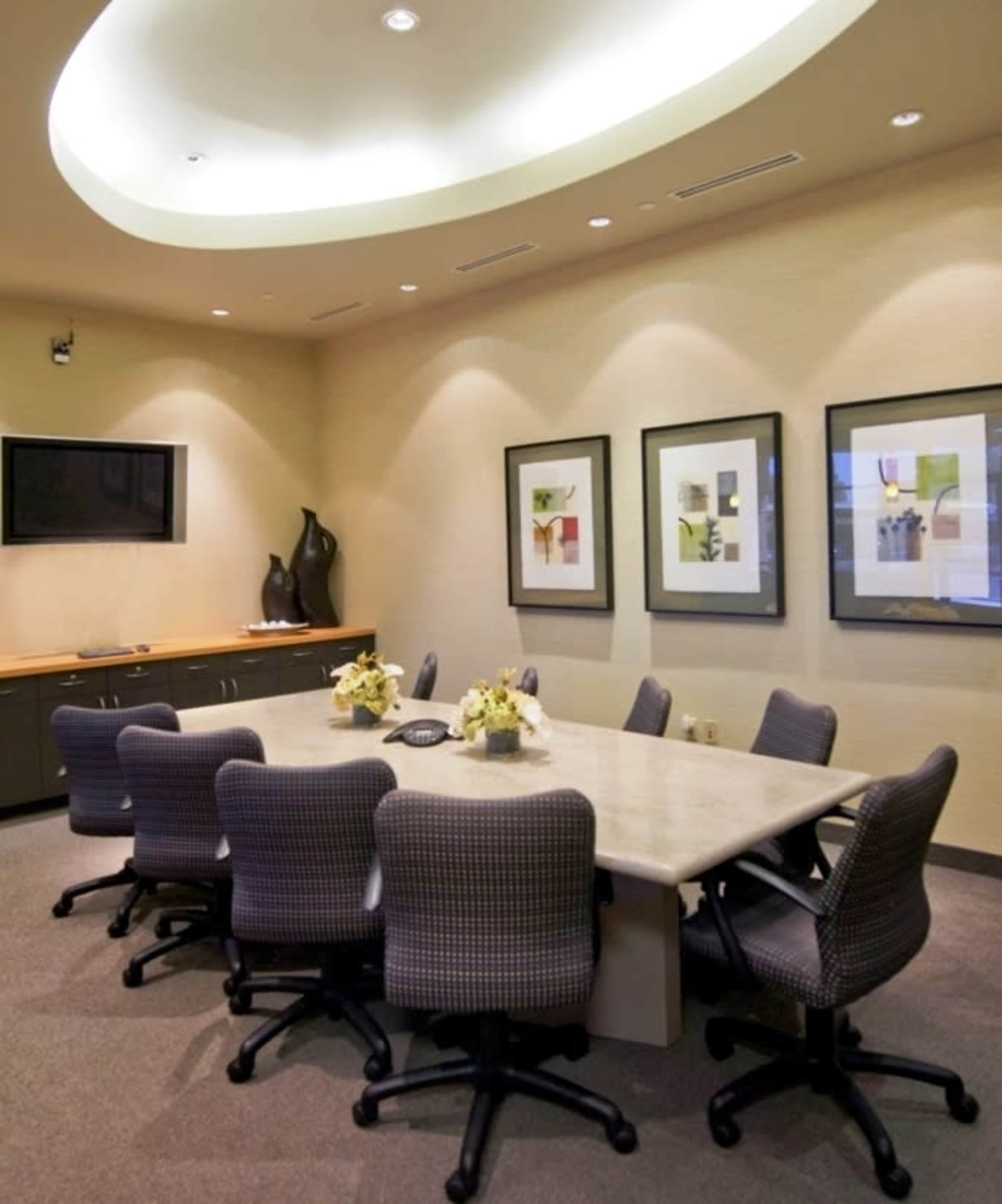 A conference room features a long table surrounded by ten chairs, with framed artwork on the walls and a television mounted in one corner.