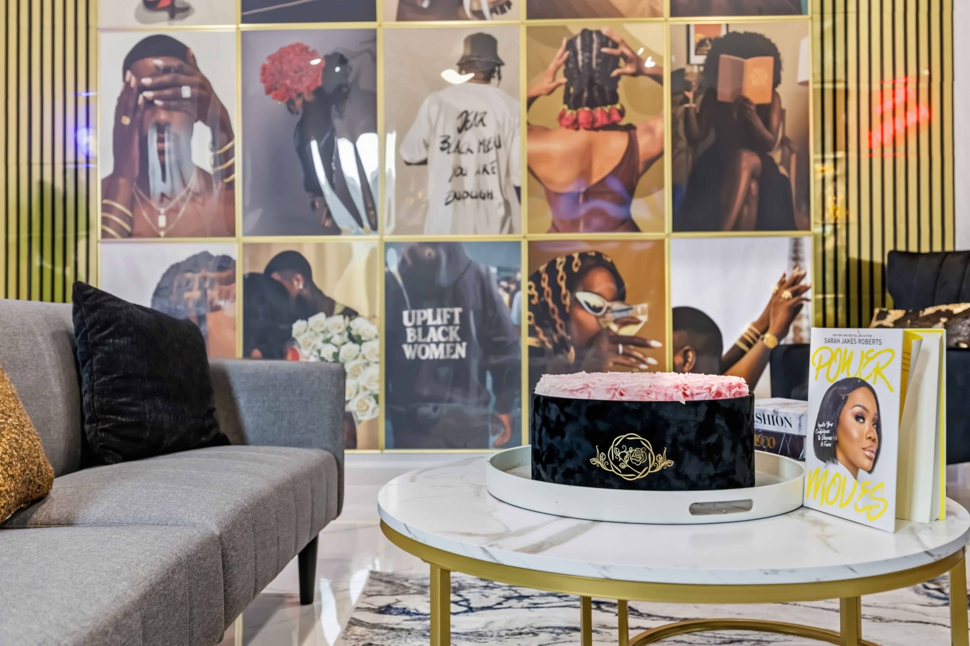 The image features a stylish living area with a marble coffee table, a black and pink cake on top, and a wall adorned with vibrant, framed photographs.