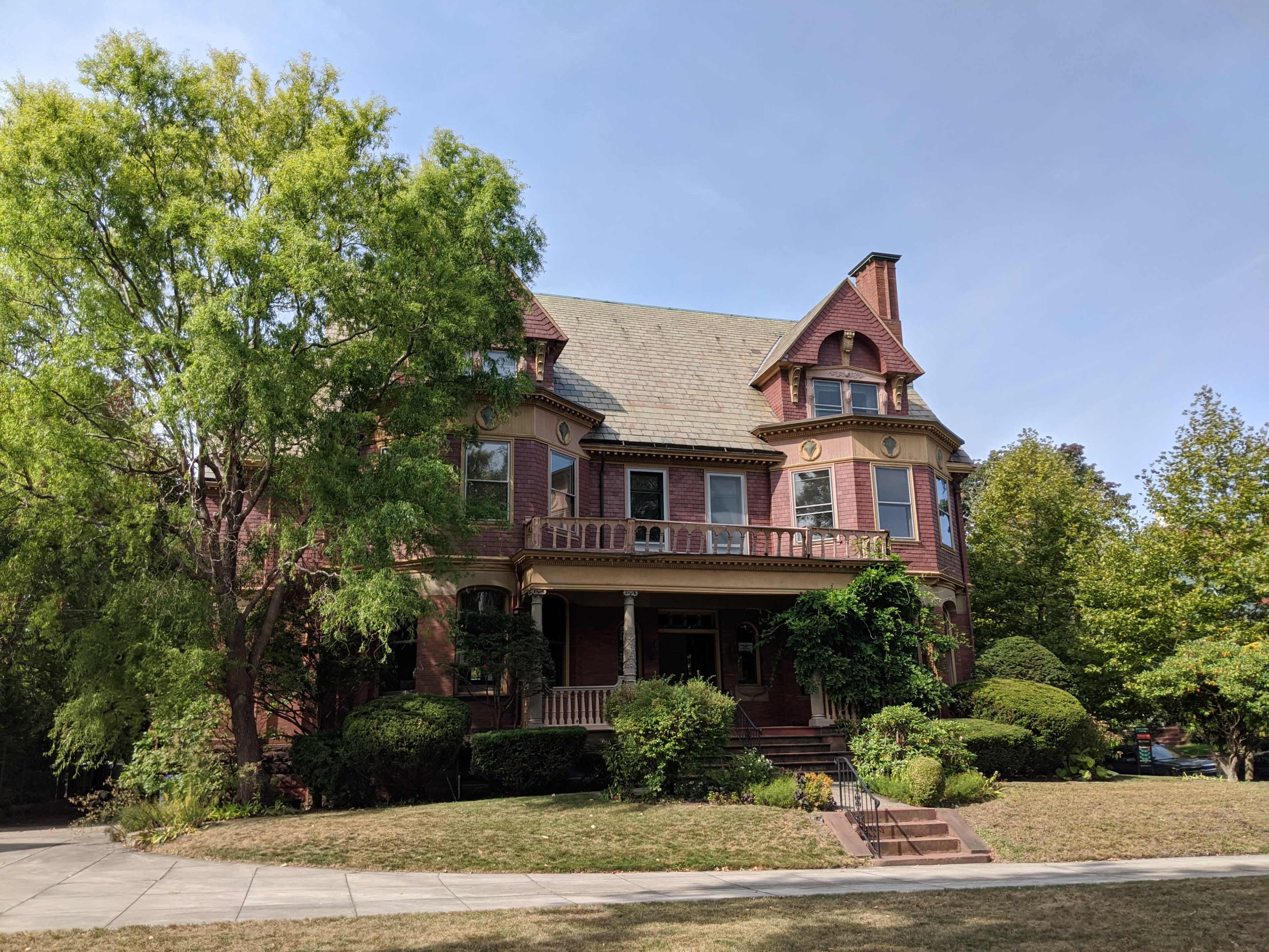 A large, red brick Victorian house stands surrounded by lush greenery and well-maintained shrubs on a clear day.
