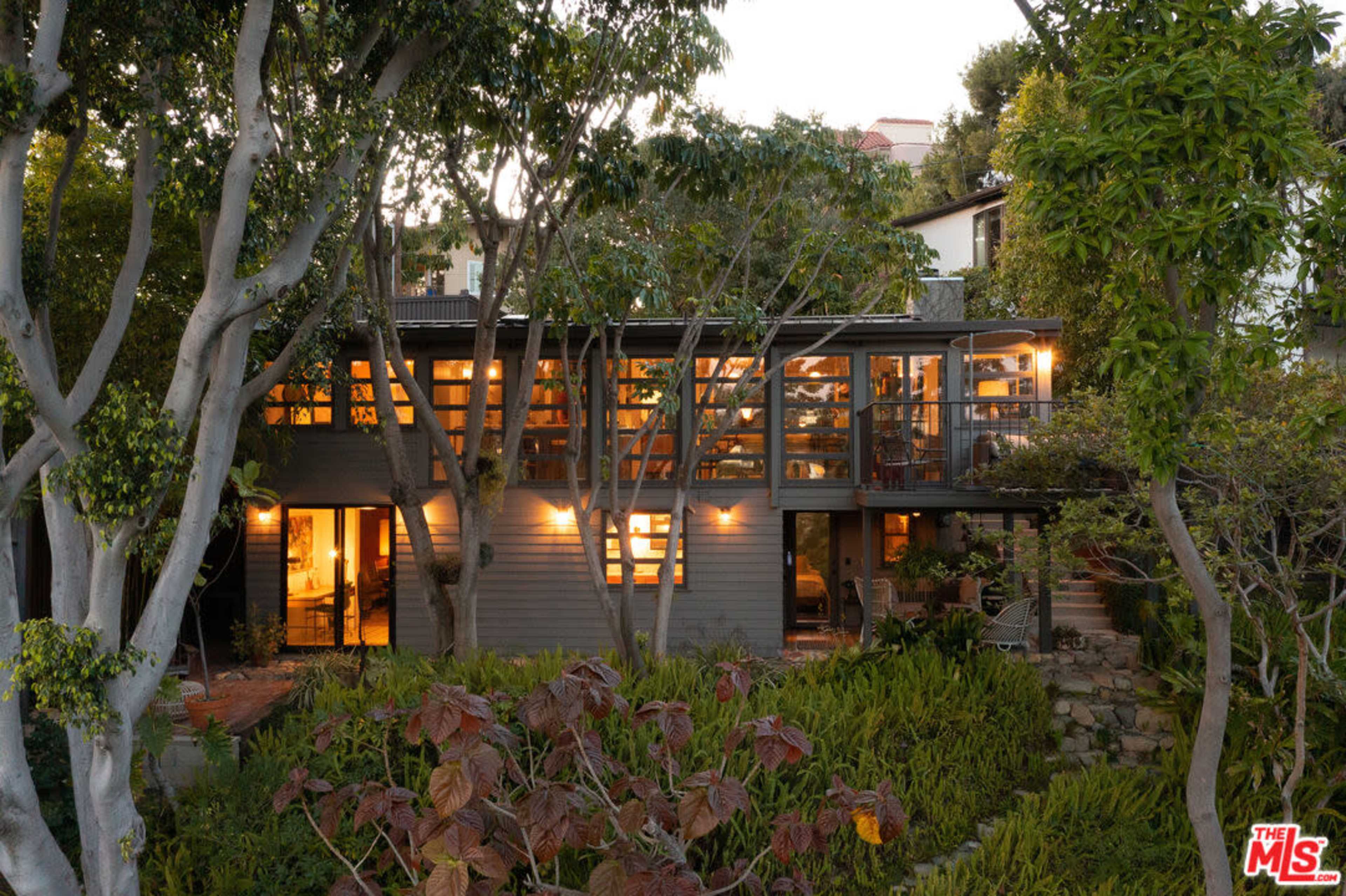A two-story, modern residential house features large windows and a deck, surrounded by landscaped greenery and trees.