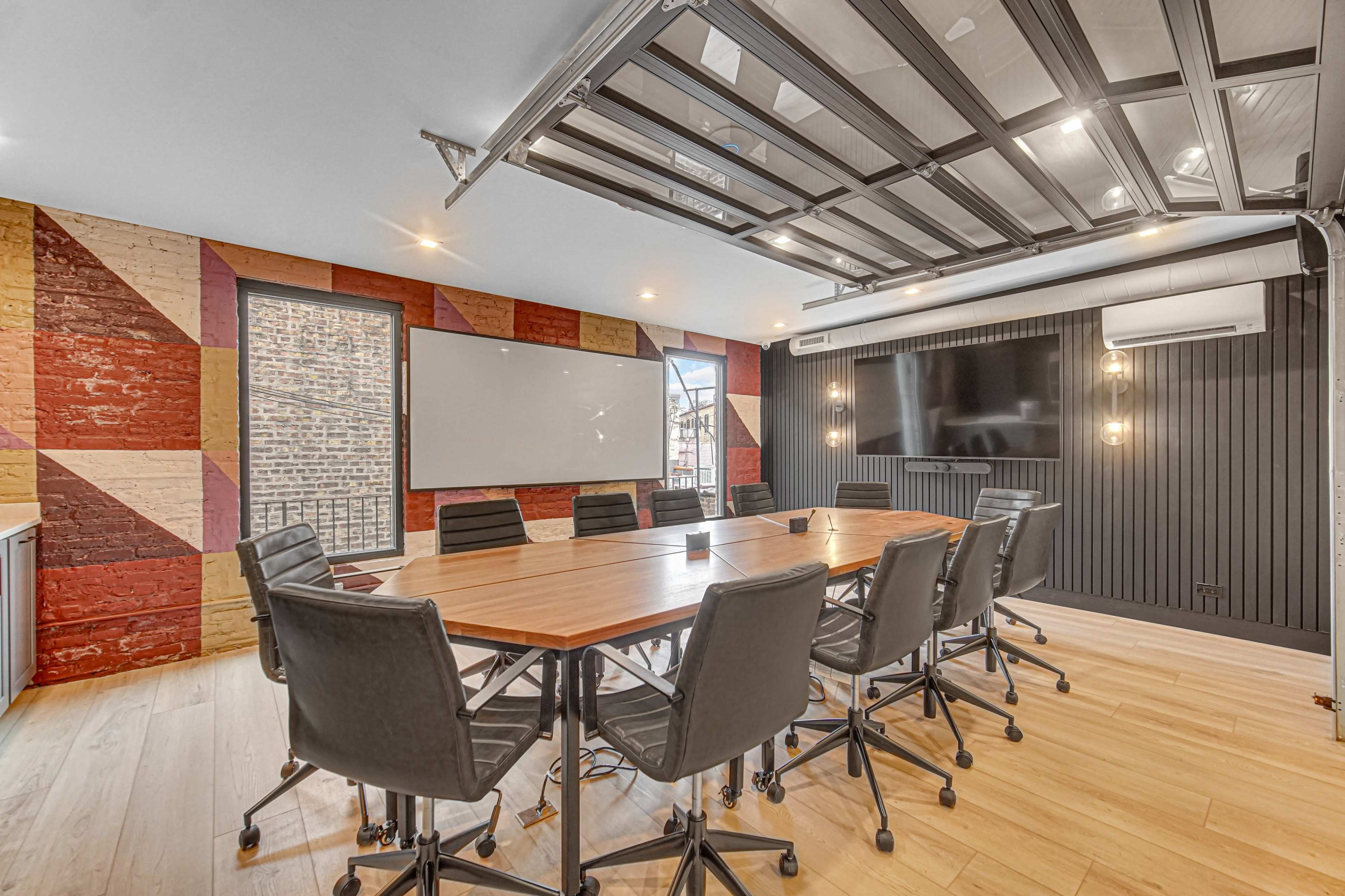The image shows a modern conference room featuring a large wooden table surrounded by black rolling chairs, with brick and black panel walls, and large windows allowing natural light.