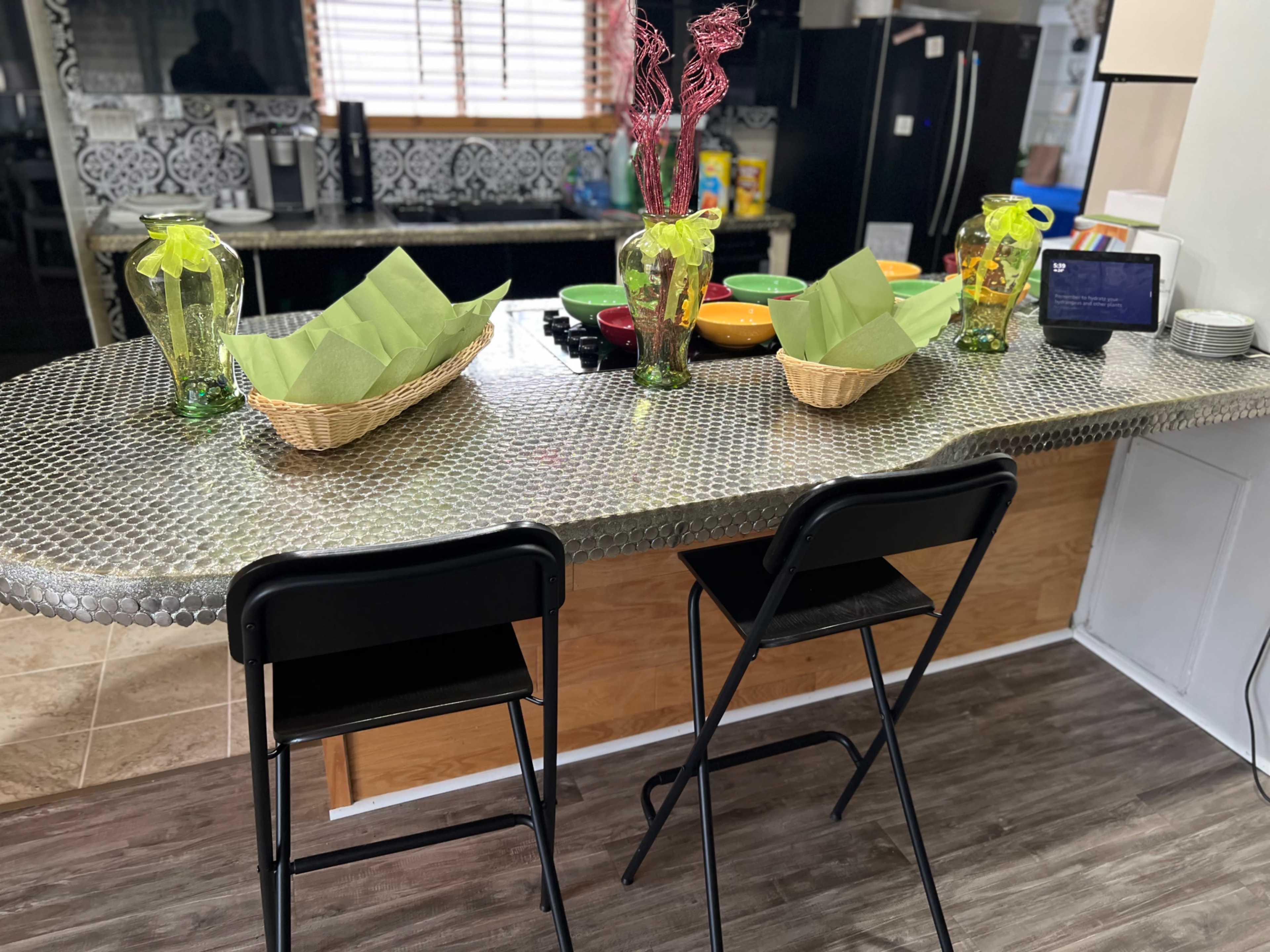 The image shows a kitchen bar area with a metallic countertop, two black stools, and decorative items including vases and baskets with green napkins.