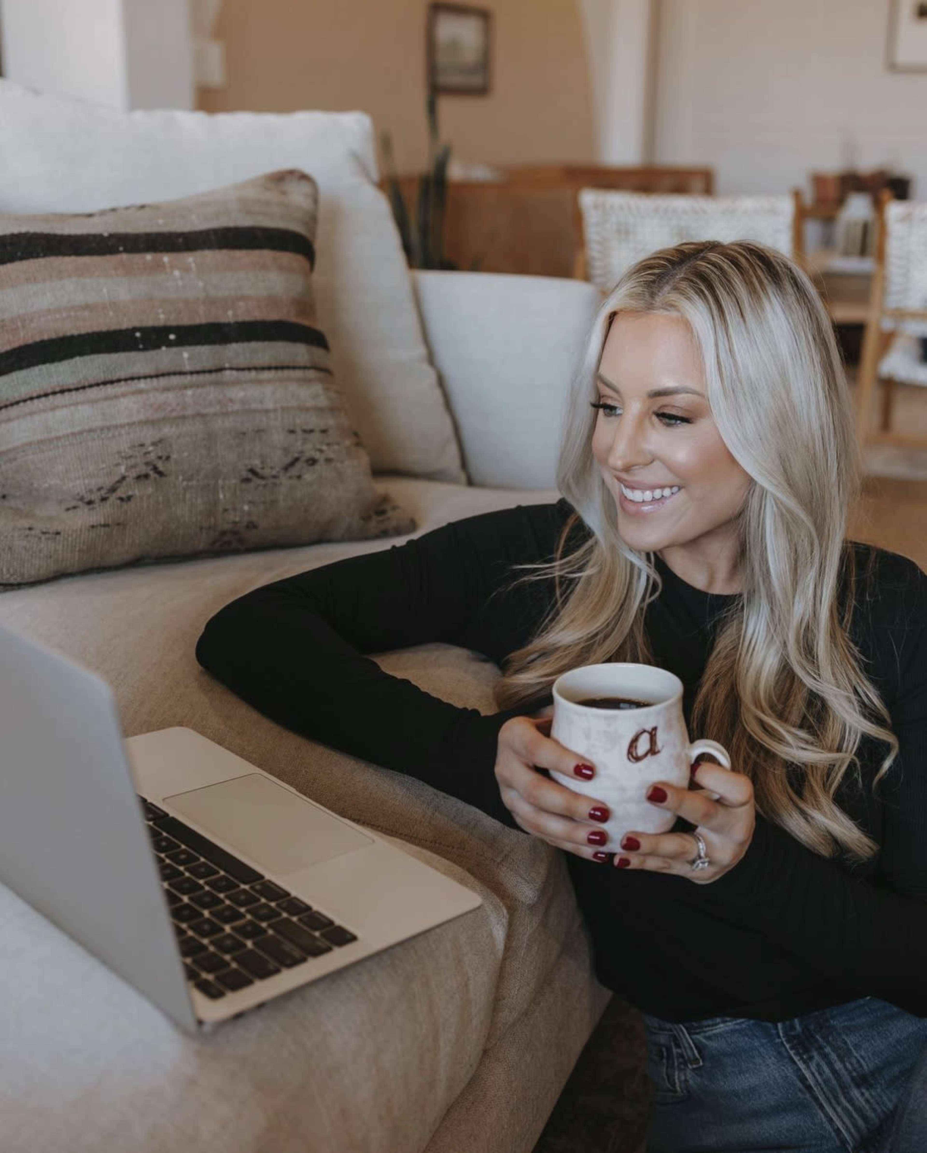 A woman sits on a sofa holding a coffee mug while looking at a laptop screen.