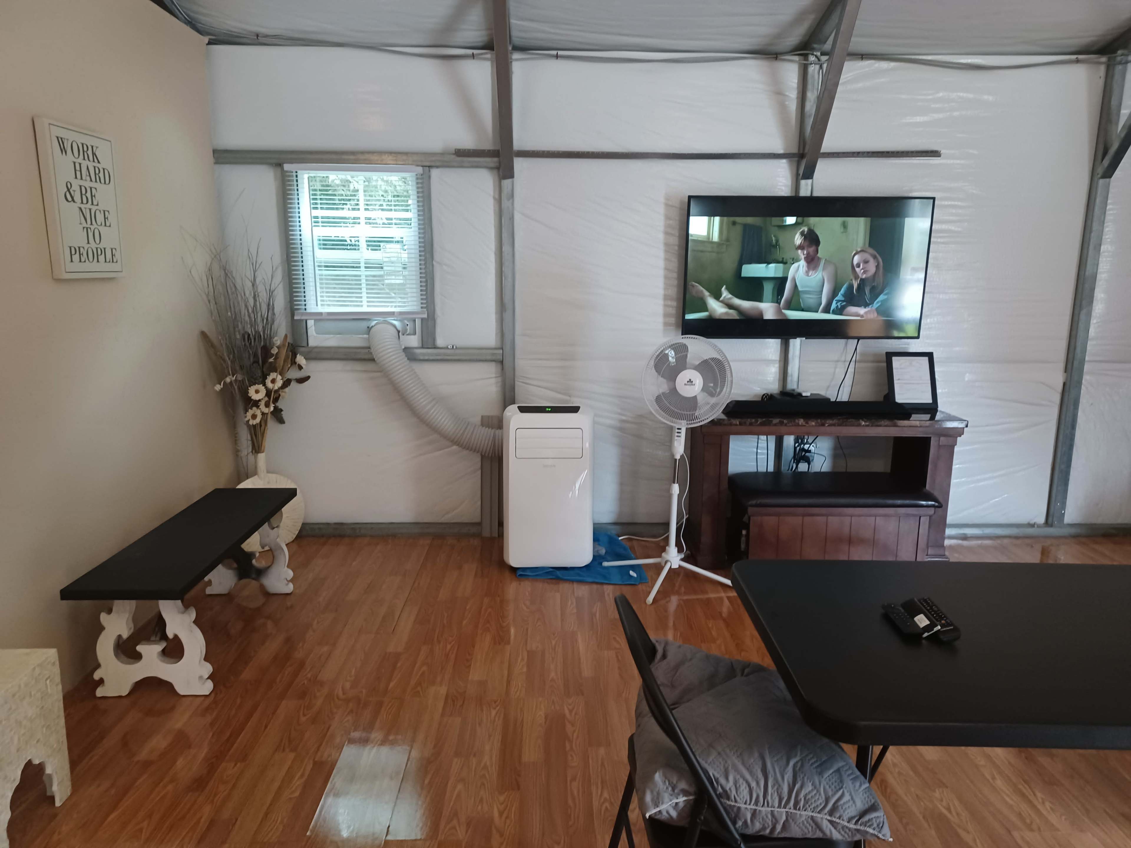 The image shows an interior of a small room with a television on a wooden stand, a fan, and a portable air conditioner, surrounded by a table and chairs.
