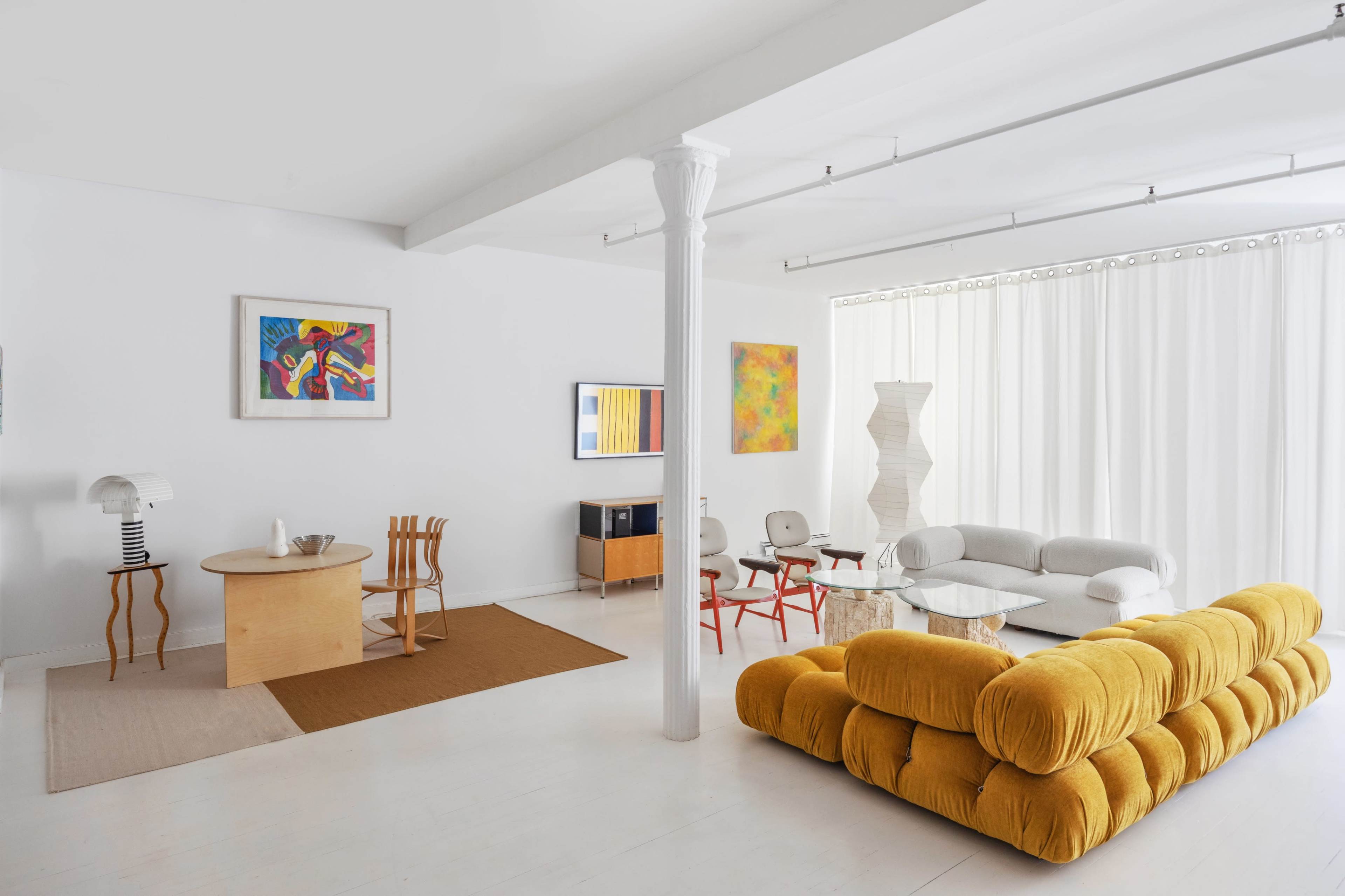 The image shows a modern living room featuring a yellow sofa, a round wooden table, various art pieces on the walls, and a mix of stylish chairs.