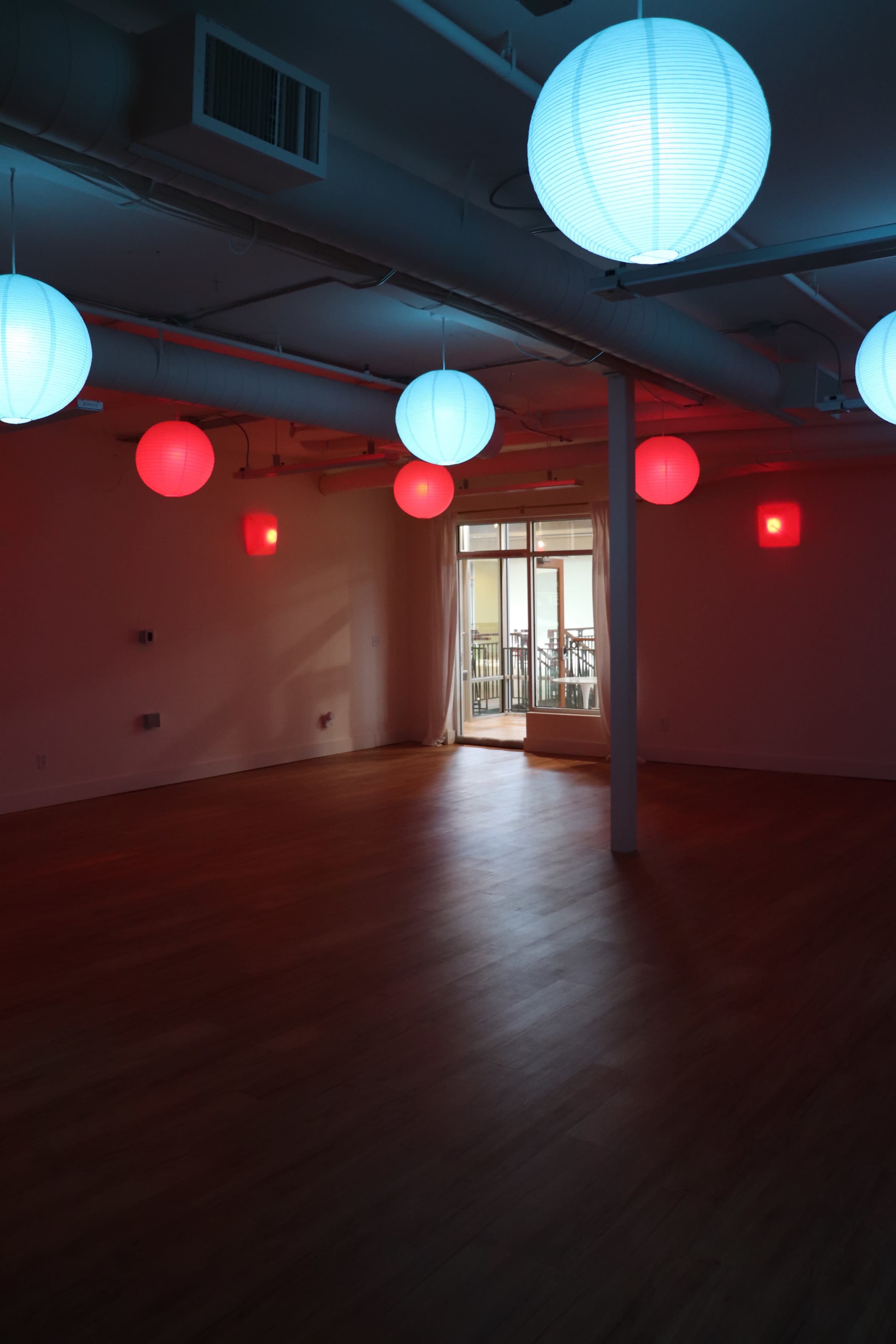 The image shows a spacious room with wooden flooring illuminated by red and blue paper lanterns hanging from the ceiling, and a doorway leading to a balcony.