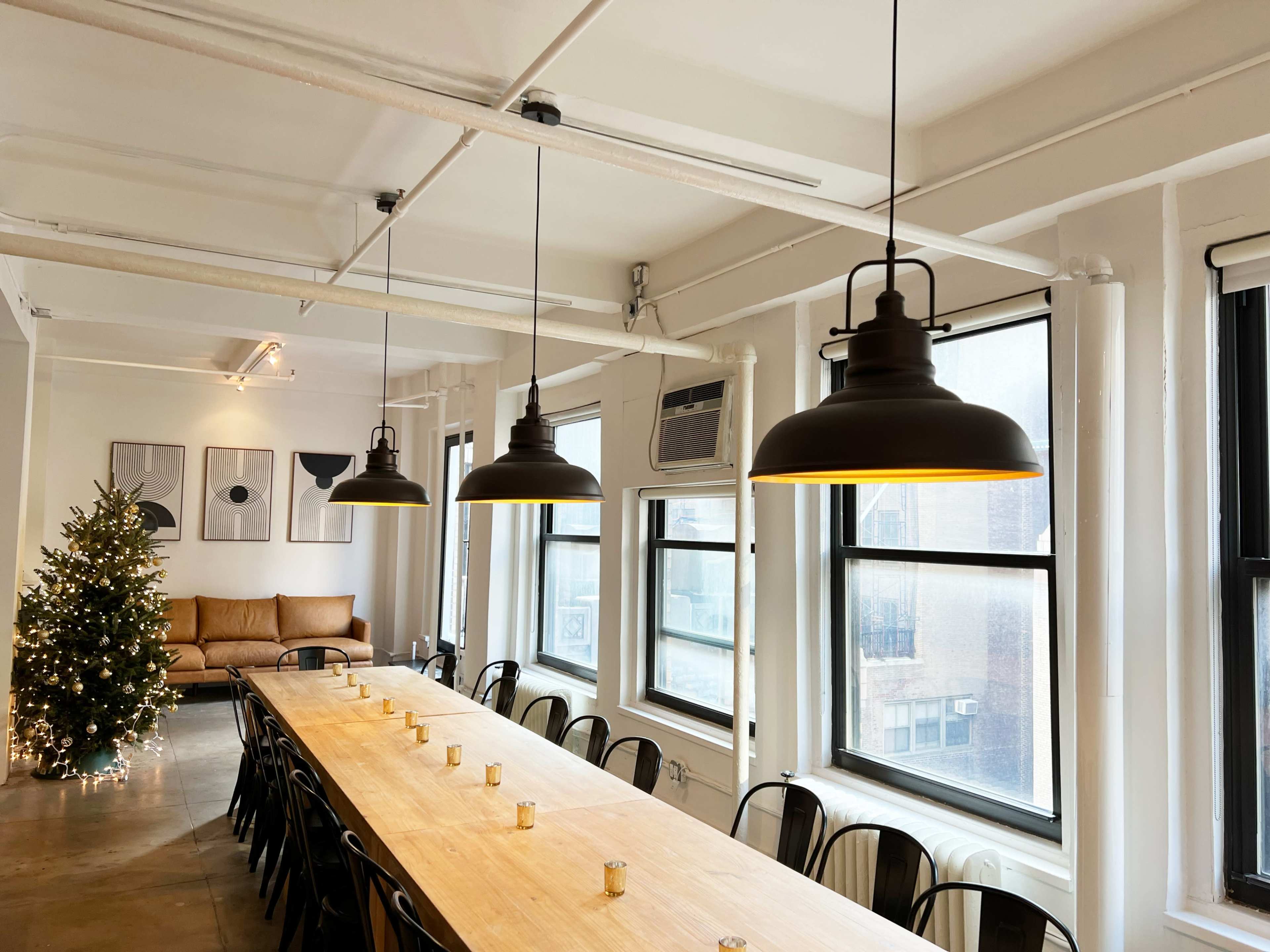 A long wooden table is set under industrial-style pendant lights in a well-lit room with windows, a couch, and a Christmas tree in the corner.