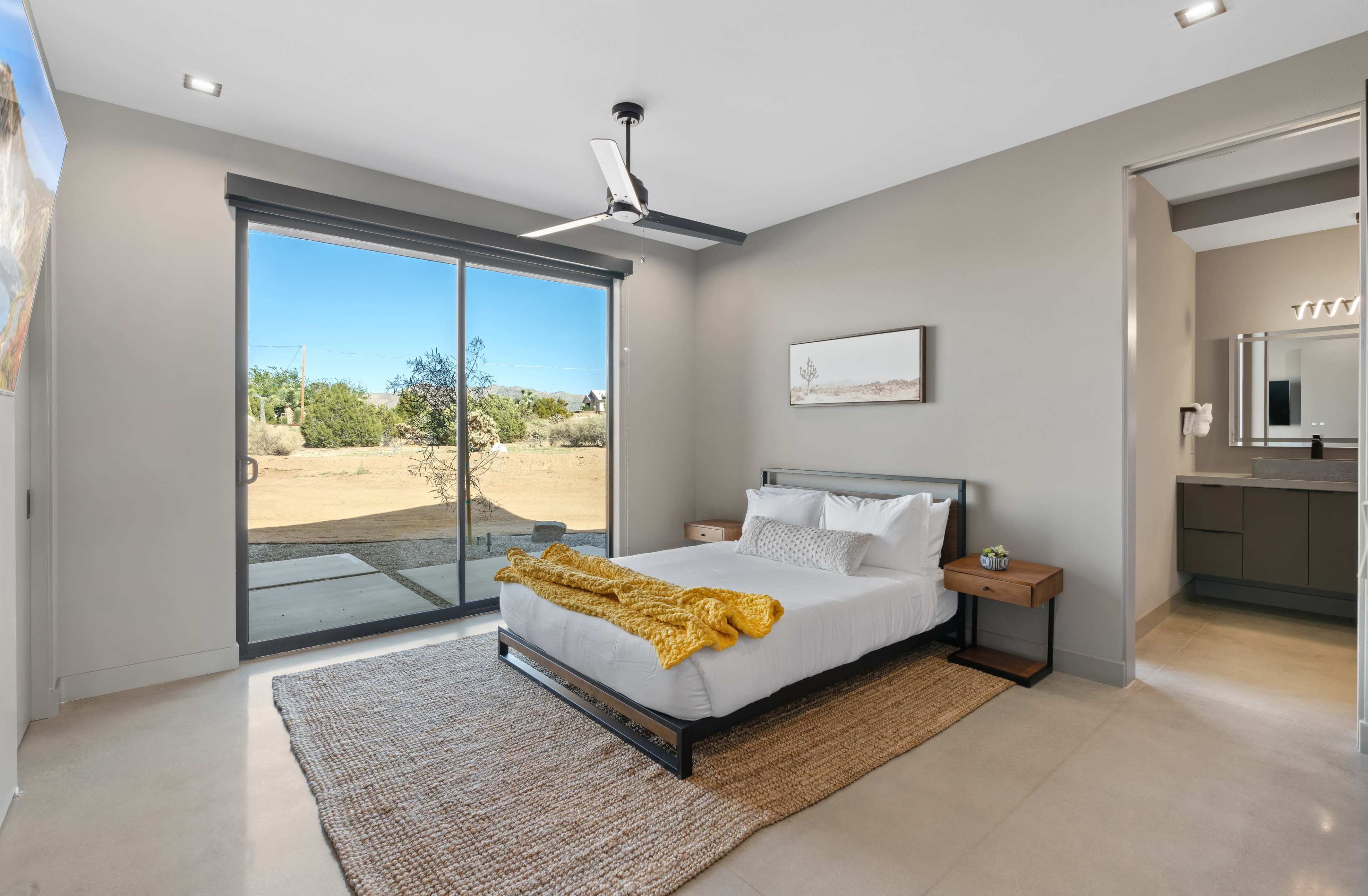 The image shows a modern bedroom featuring a bed with white bedding, a wooden nightstand, and large glass sliding doors leading to an outdoor area.