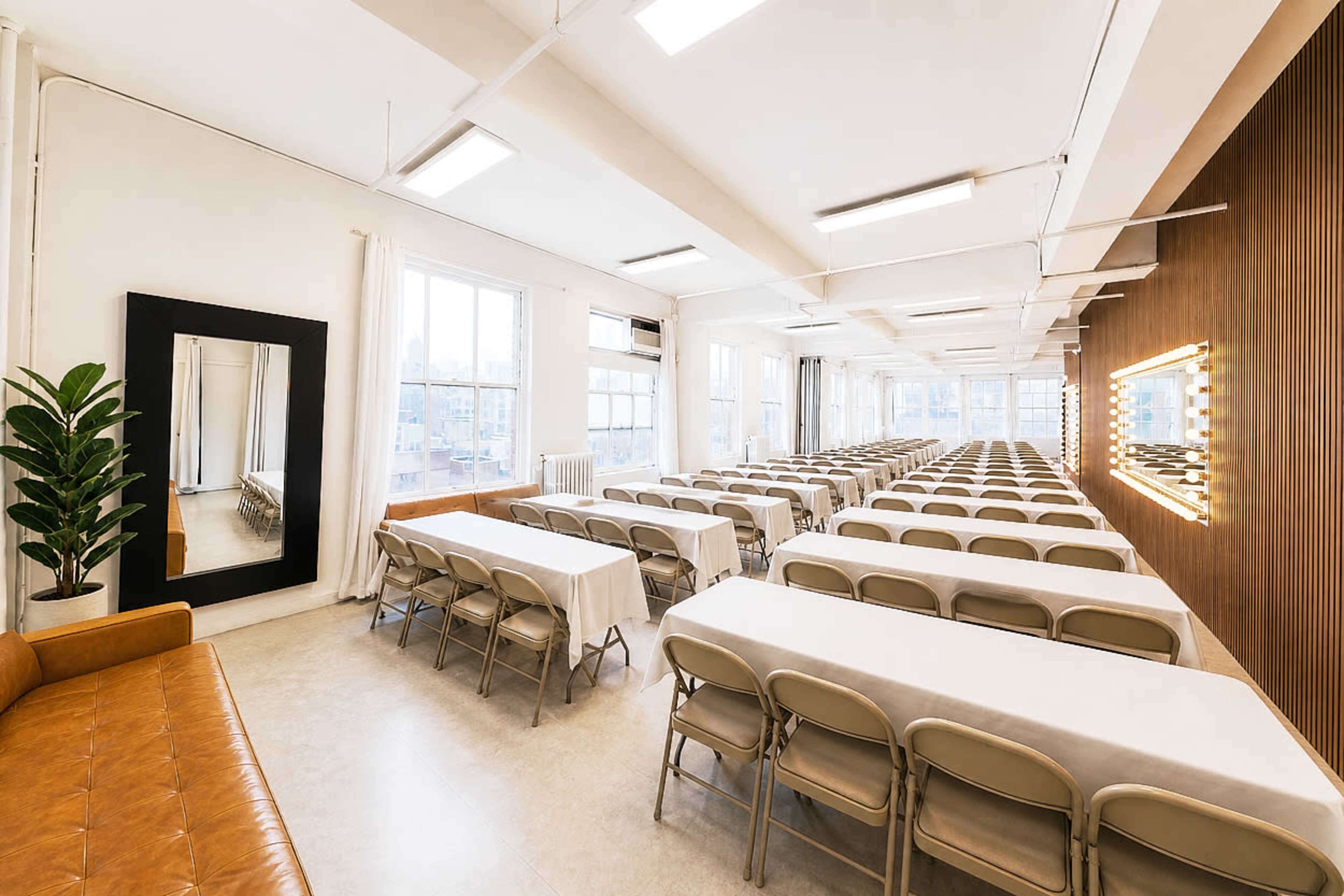 The image shows a spacious room arranged with several long tables and chairs, suitable for an event or meeting, with large windows allowing natural light to enter.