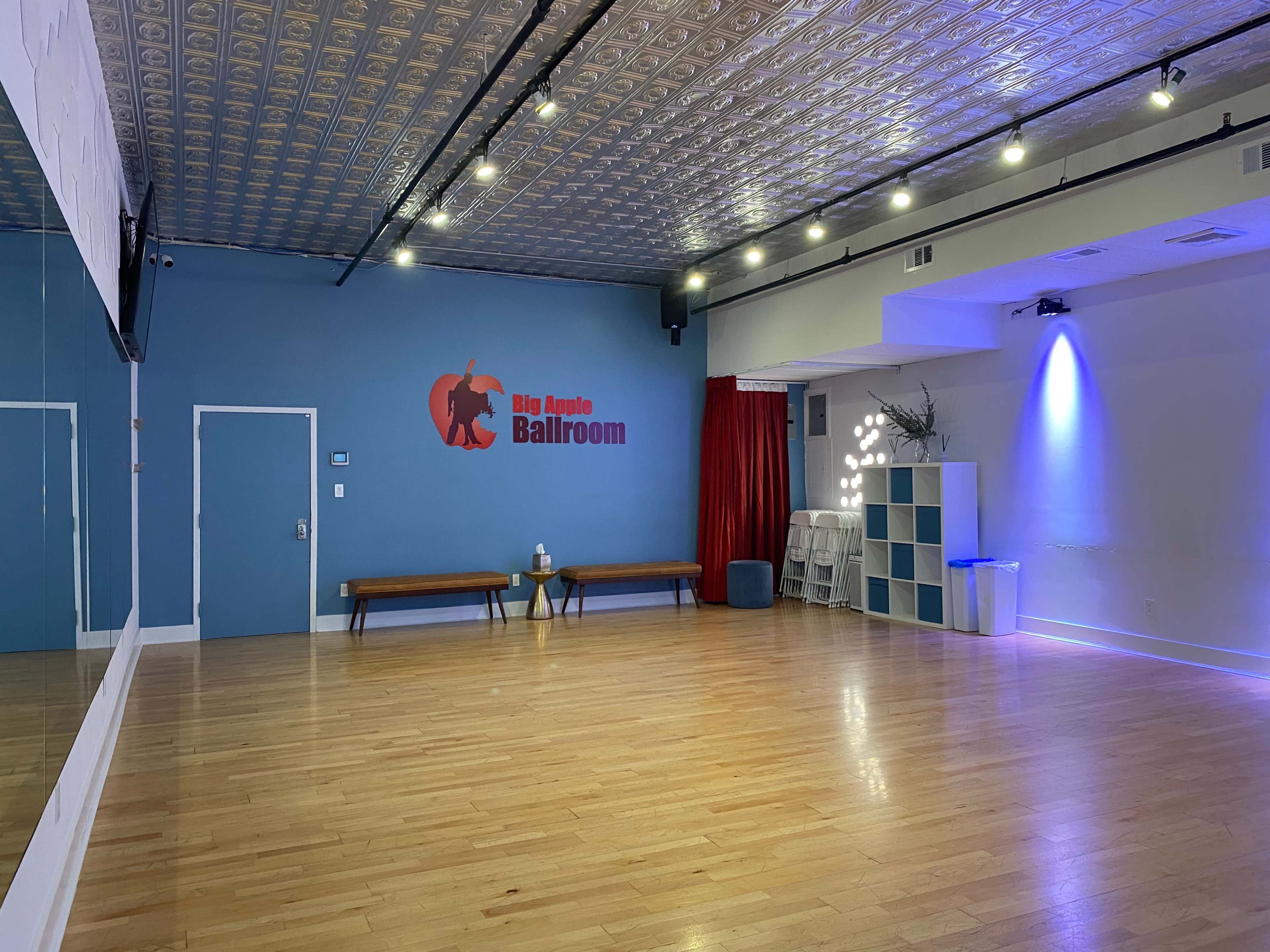 The image shows an empty ballroom with a wooden floor, a large mirror on one wall, and a red curtain along another wall, featuring the logo "Big Apple Ballroom" on the blue painted wall.
