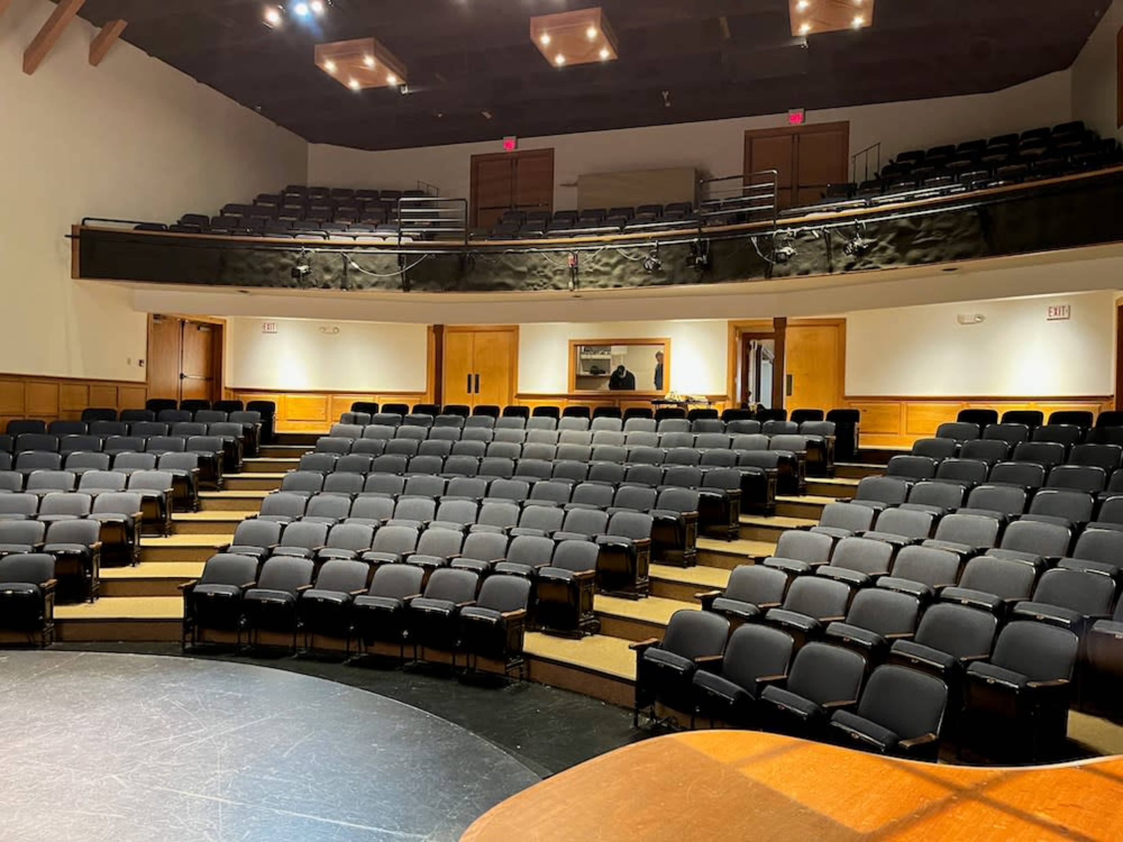 A theater with tiered seating and a circular stage at the front, empty rows of black chairs visible in an intimate setting.