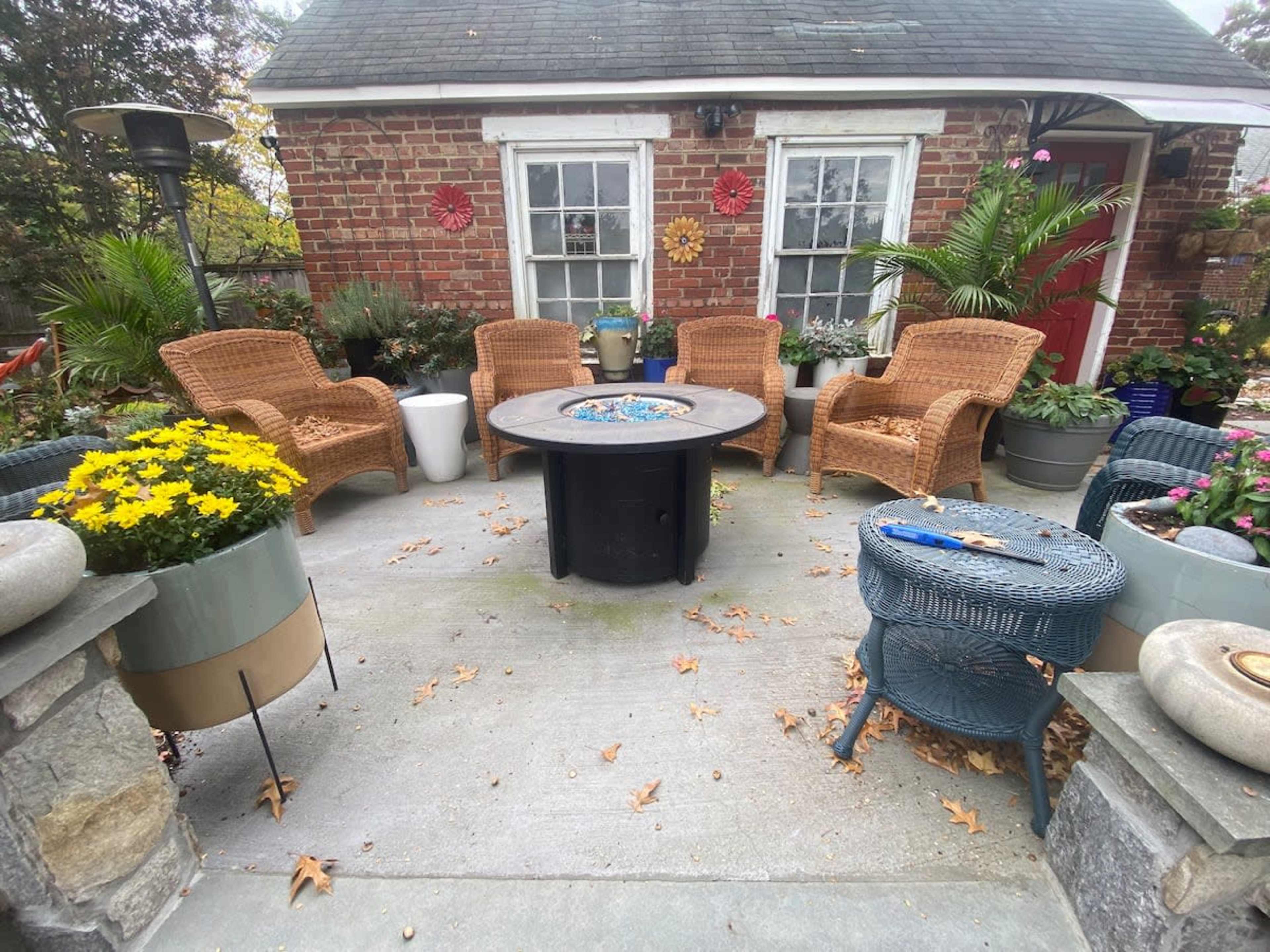 A patio with wicker chairs arranged around a circular fire pit, surrounded by potted plants and flowers.