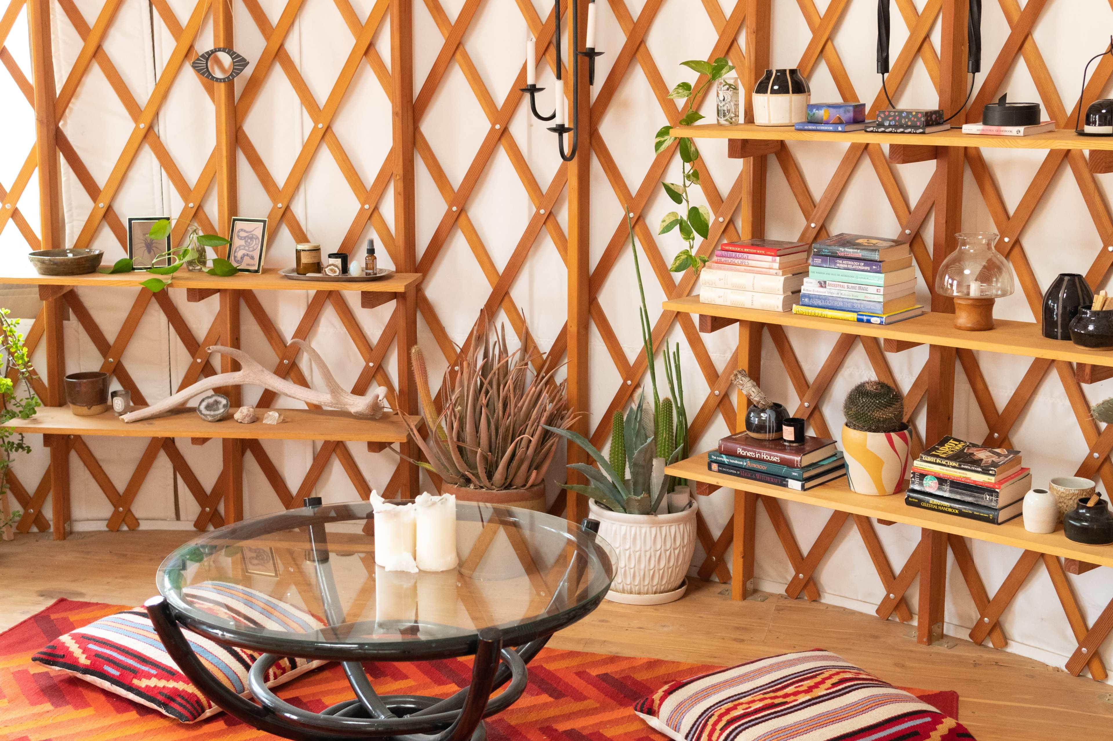 The image shows a cozy interior of a yurt featuring wooden lattice walls, shelves filled with decorative items and books, a round glass coffee table, and colorful rugs.