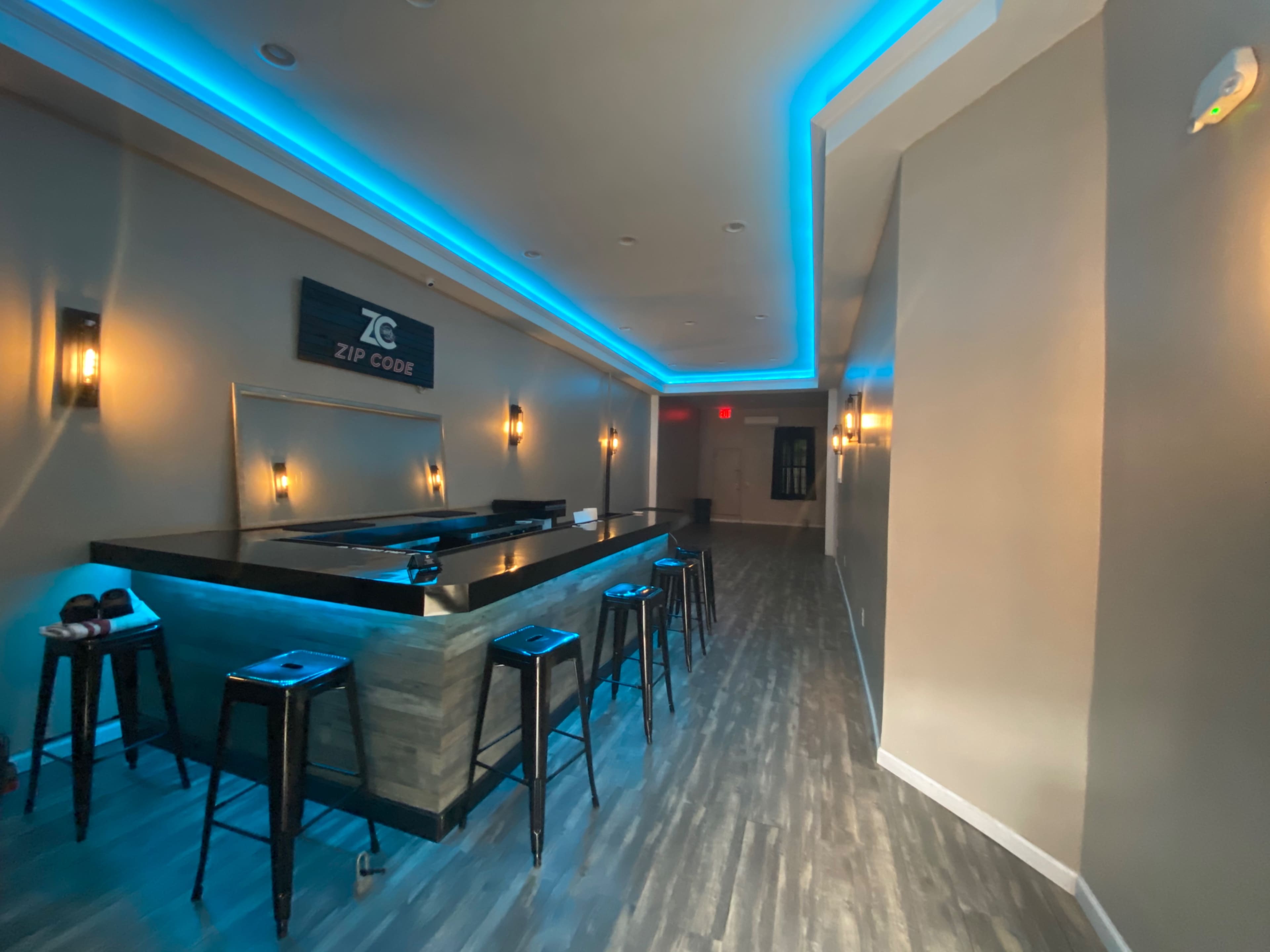 A modern bar area with a long countertop and several high stools, illuminated by blue LED lighting along the ceiling.
