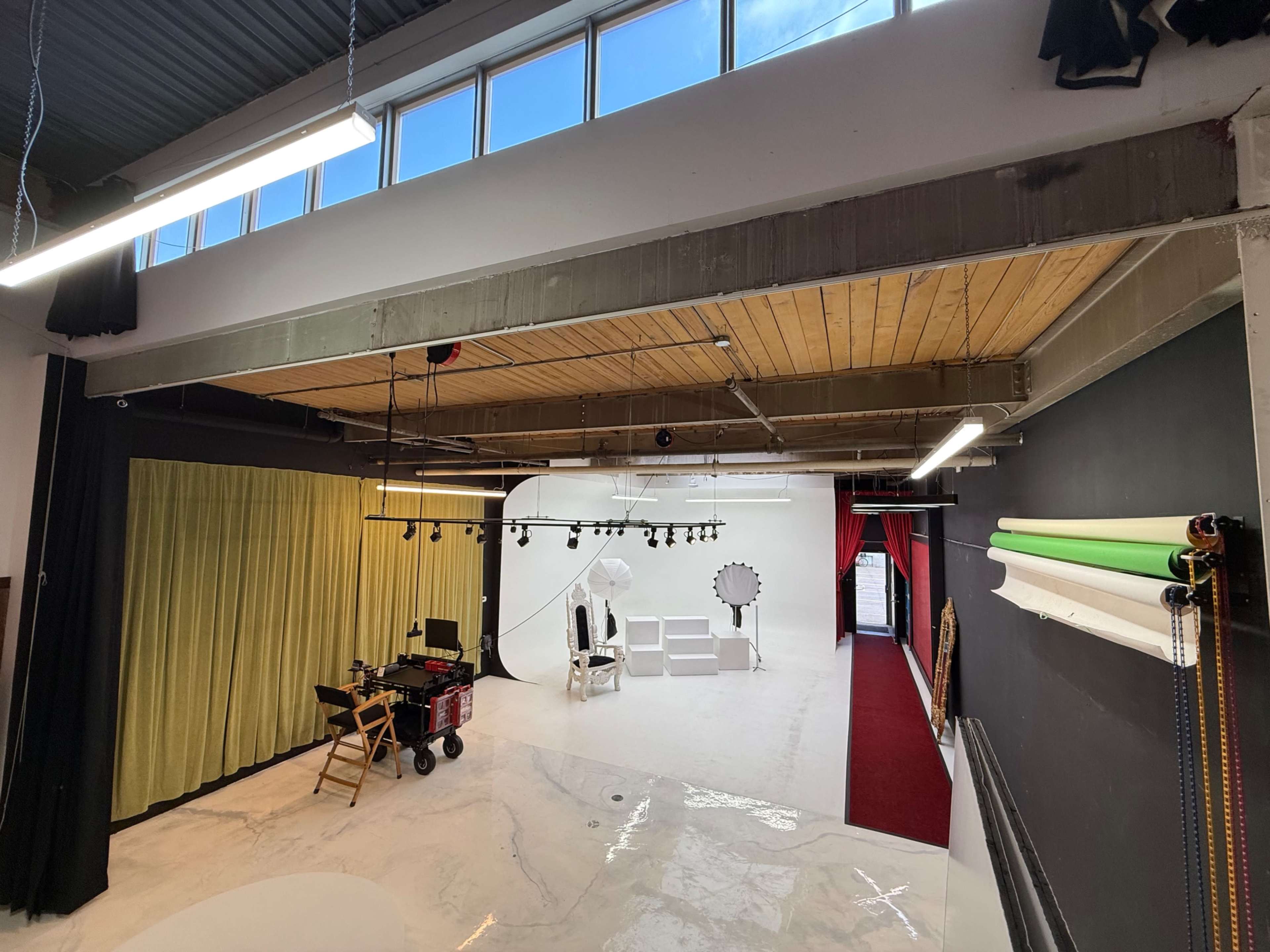 The image shows a photography studio with a white backdrop, seating area, lighting equipment, and a corridor leading to another space.