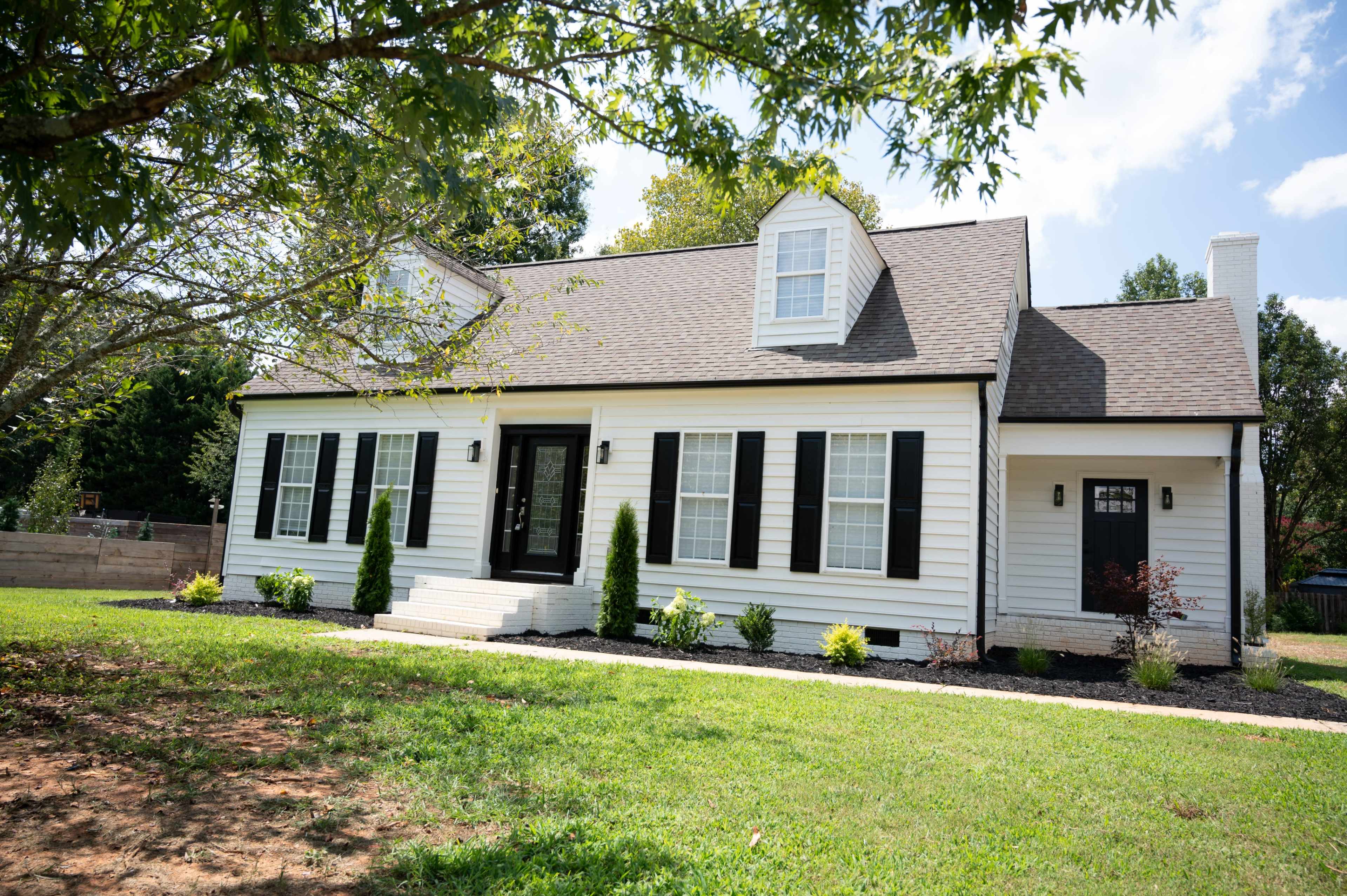 A white, single-story house with black shutters and a front porch is set on a grassy lawn surrounded by trees.