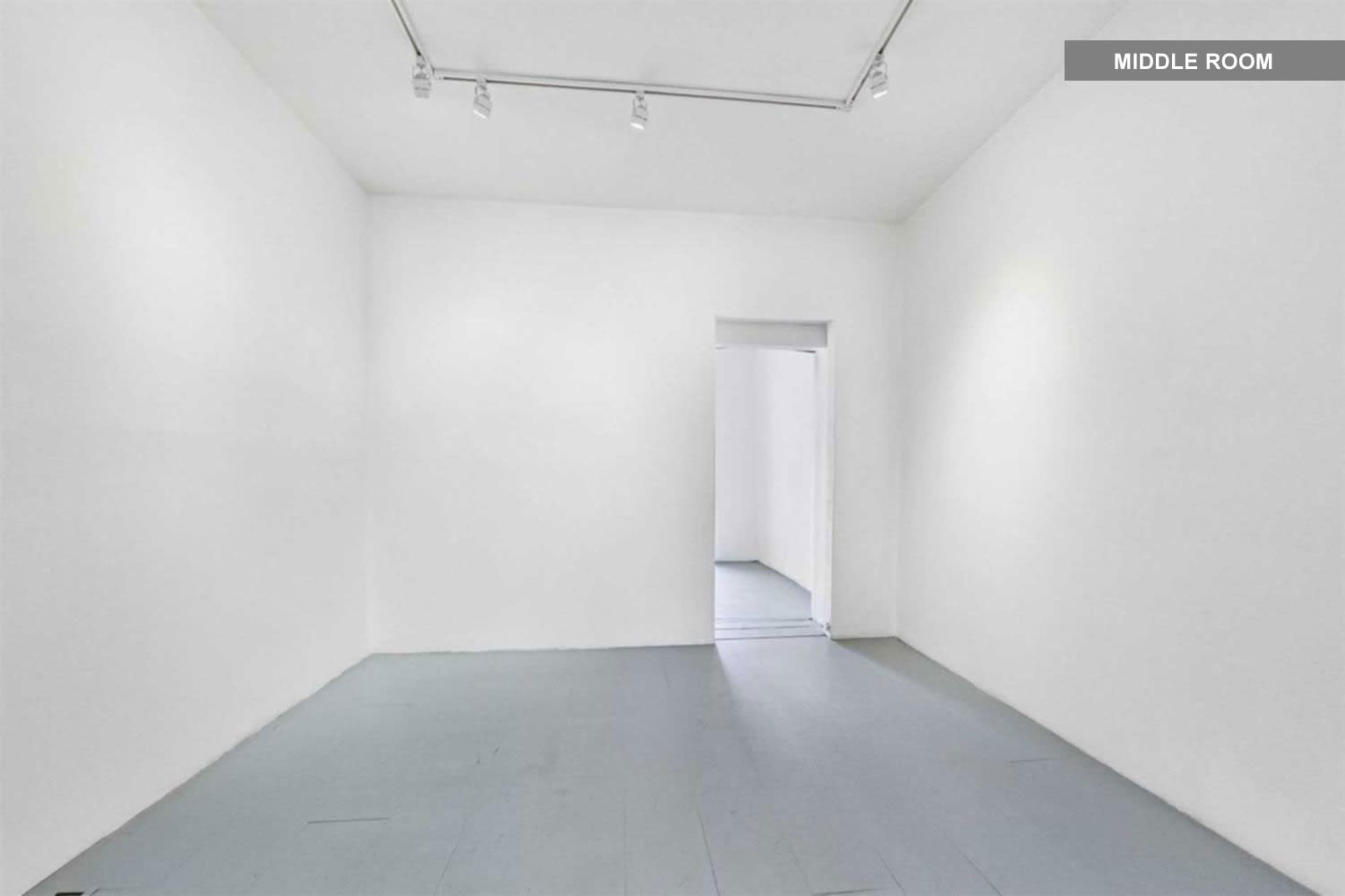 A minimalist, empty room with white walls and a light gray floor, featuring an open doorway leading to another area.