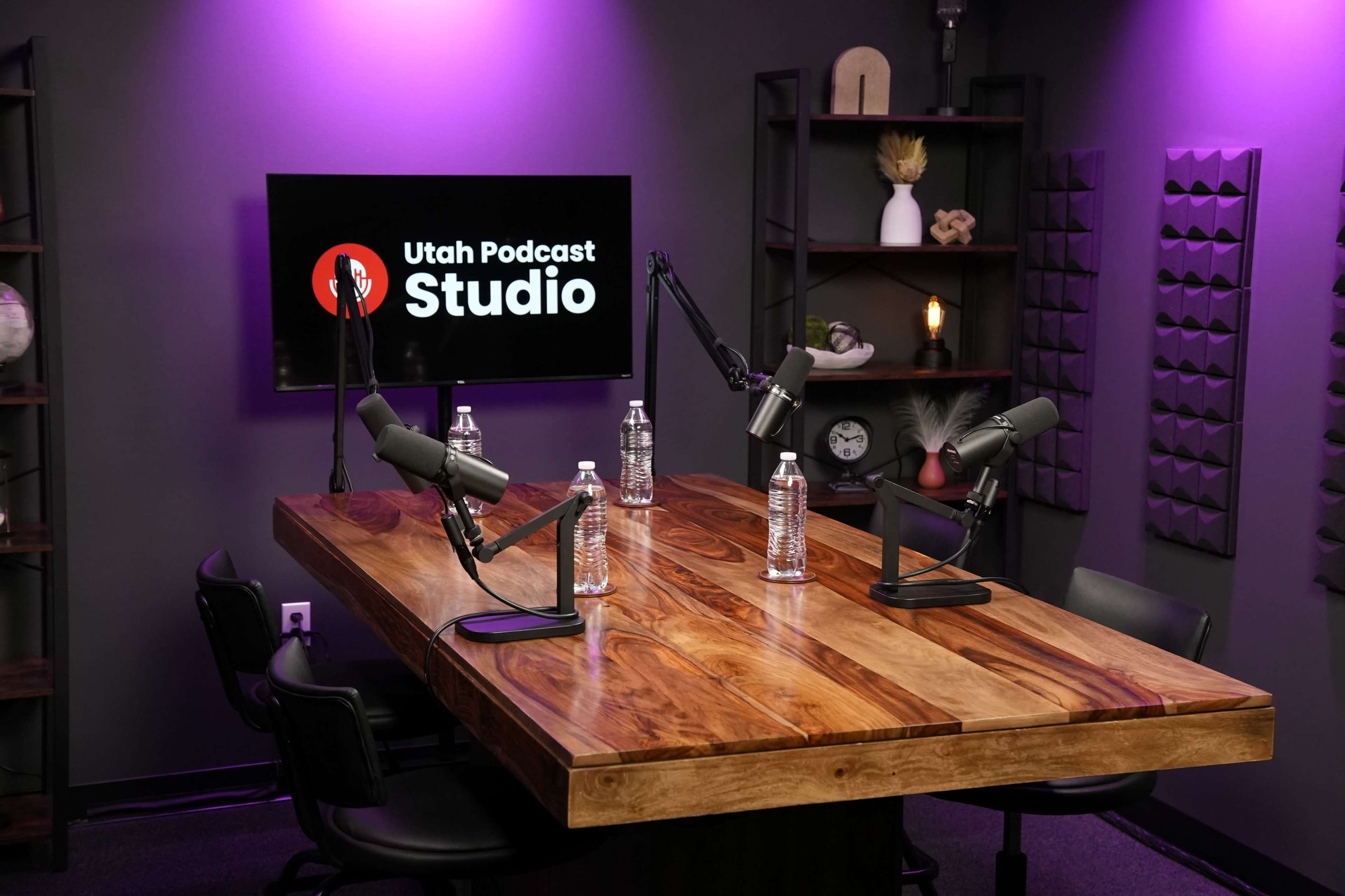 A podcast recording studio features a large wooden table with microphones and chairs, illuminated by purple lighting and displaying a "Utah Podcast Studio" logo on a screen.