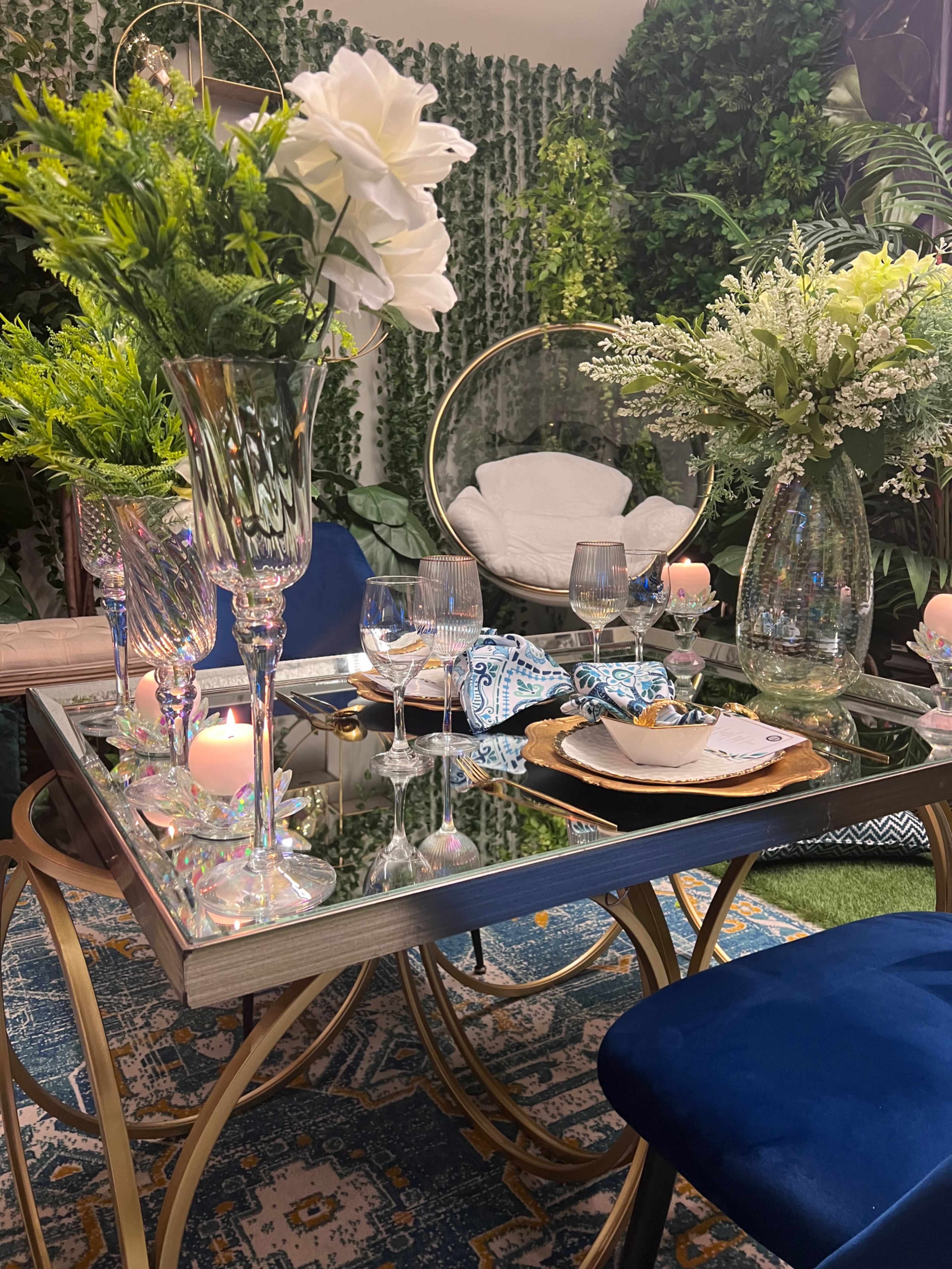 A table is set with elegant glassware and floral arrangements, surrounded by decorative greenery and plush seating.