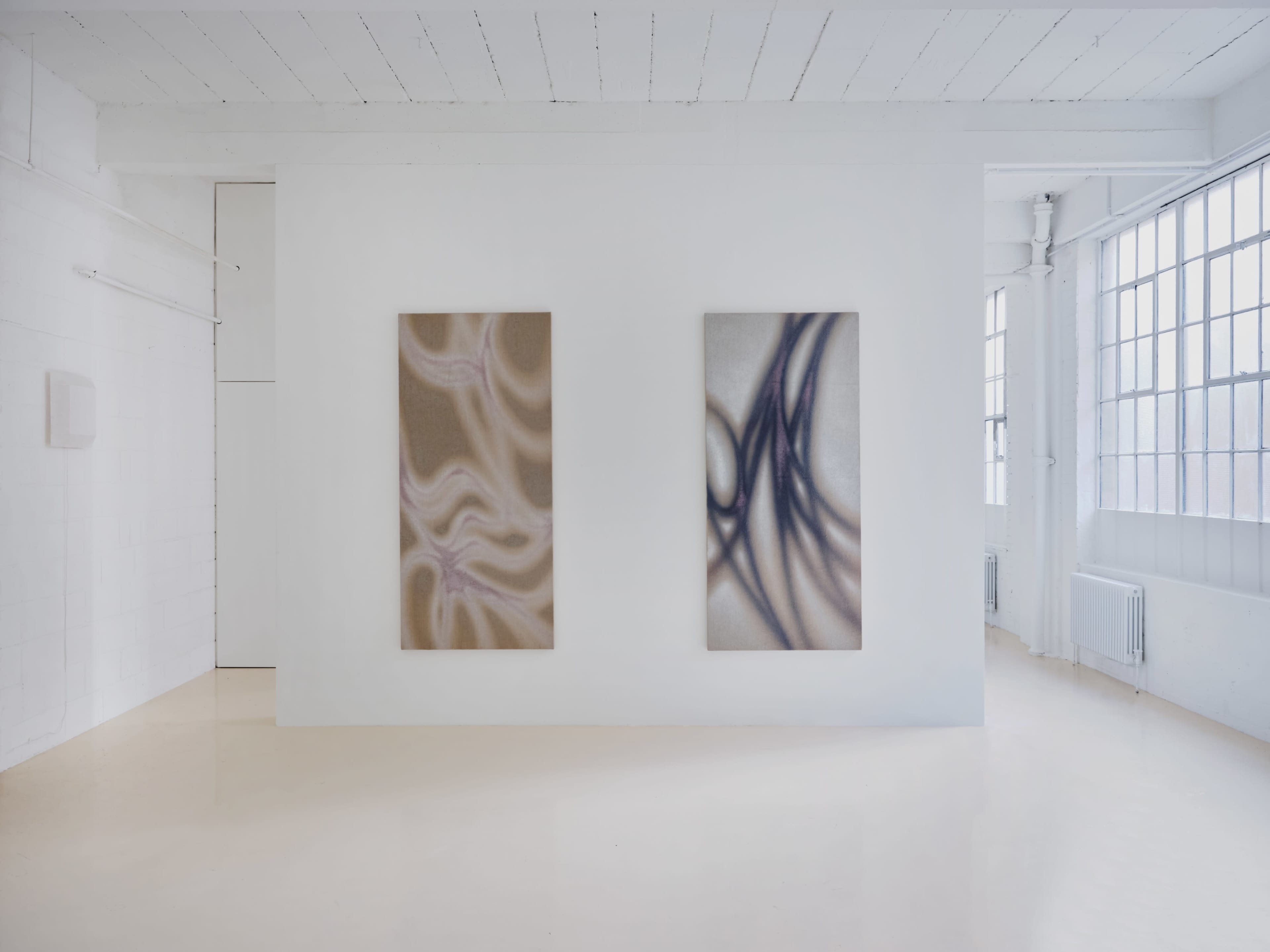 The image shows an art gallery with two large abstract paintings displayed on a white wall, illuminated by natural light from large windows.