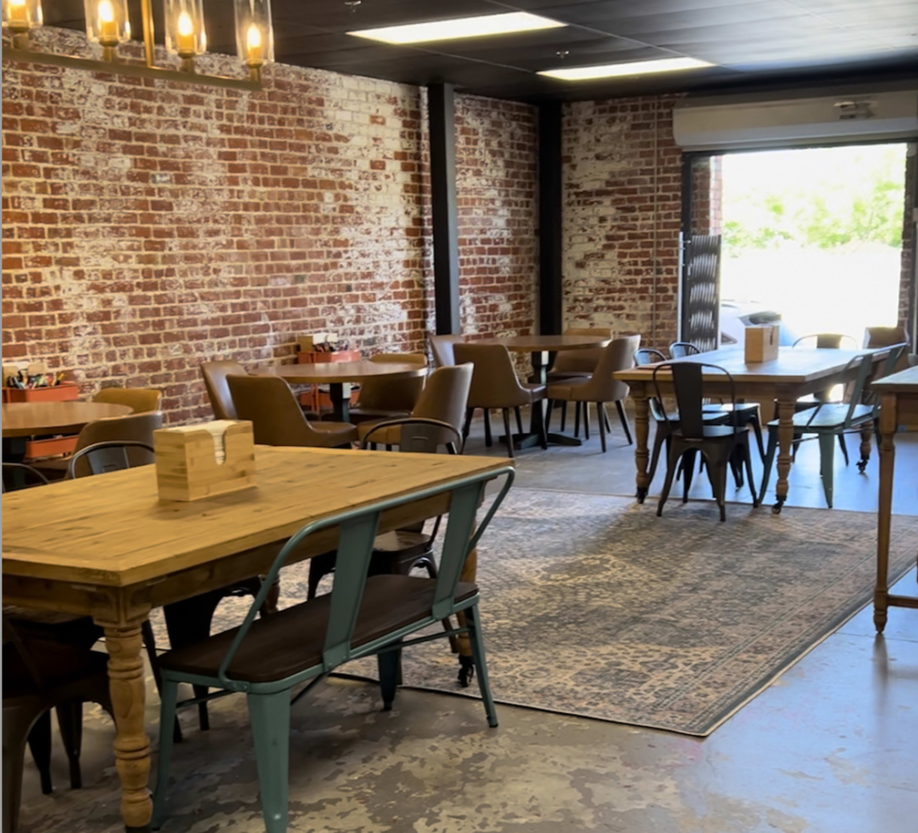 A spacious dining area with wooden tables and brown chairs, featuring exposed brick walls and a large open door leading outside.