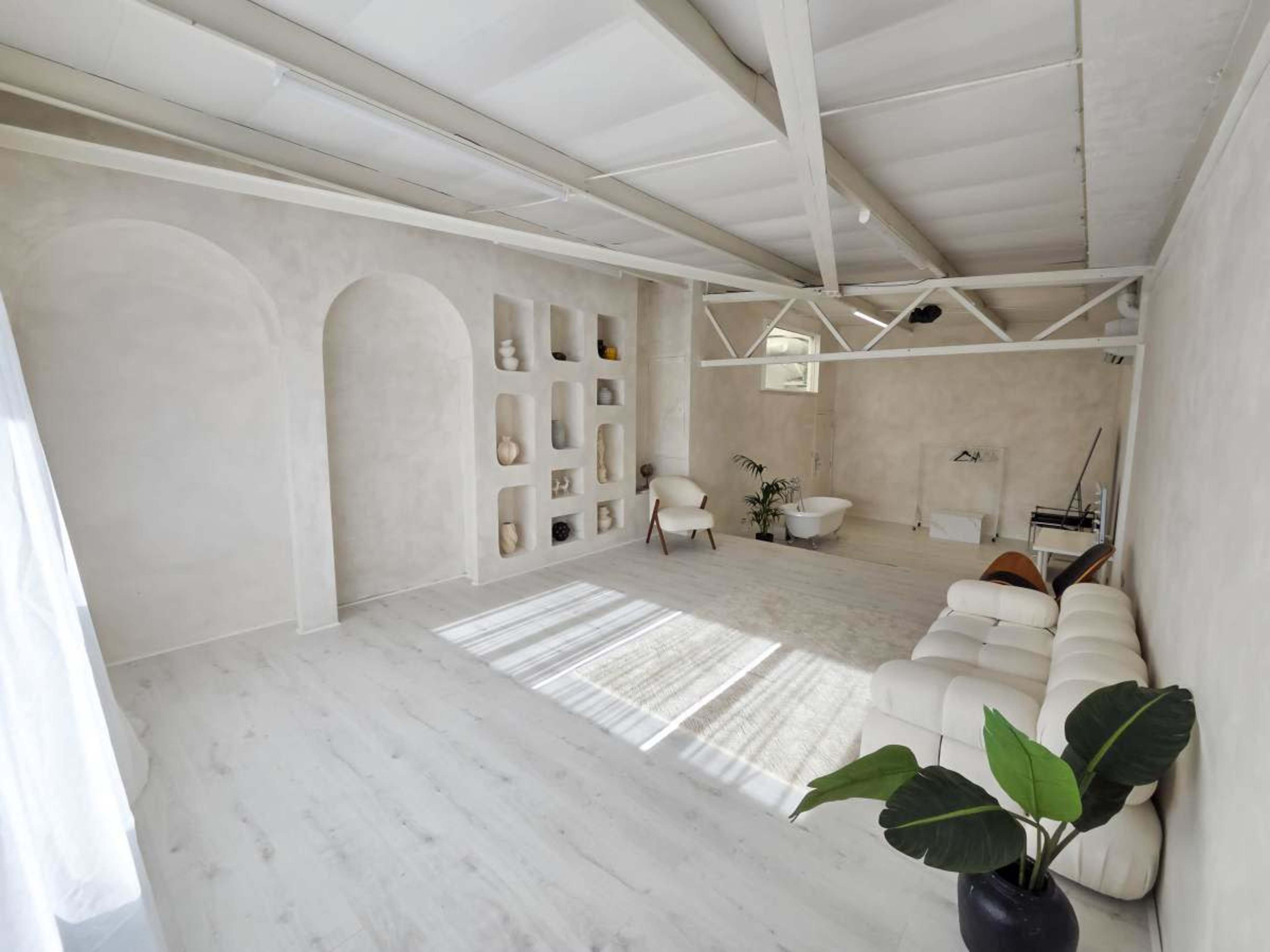 The image depicts a minimalistic interior space with light-colored walls, wooden beams, and a white sofa, featuring decorative niches and a small plant.