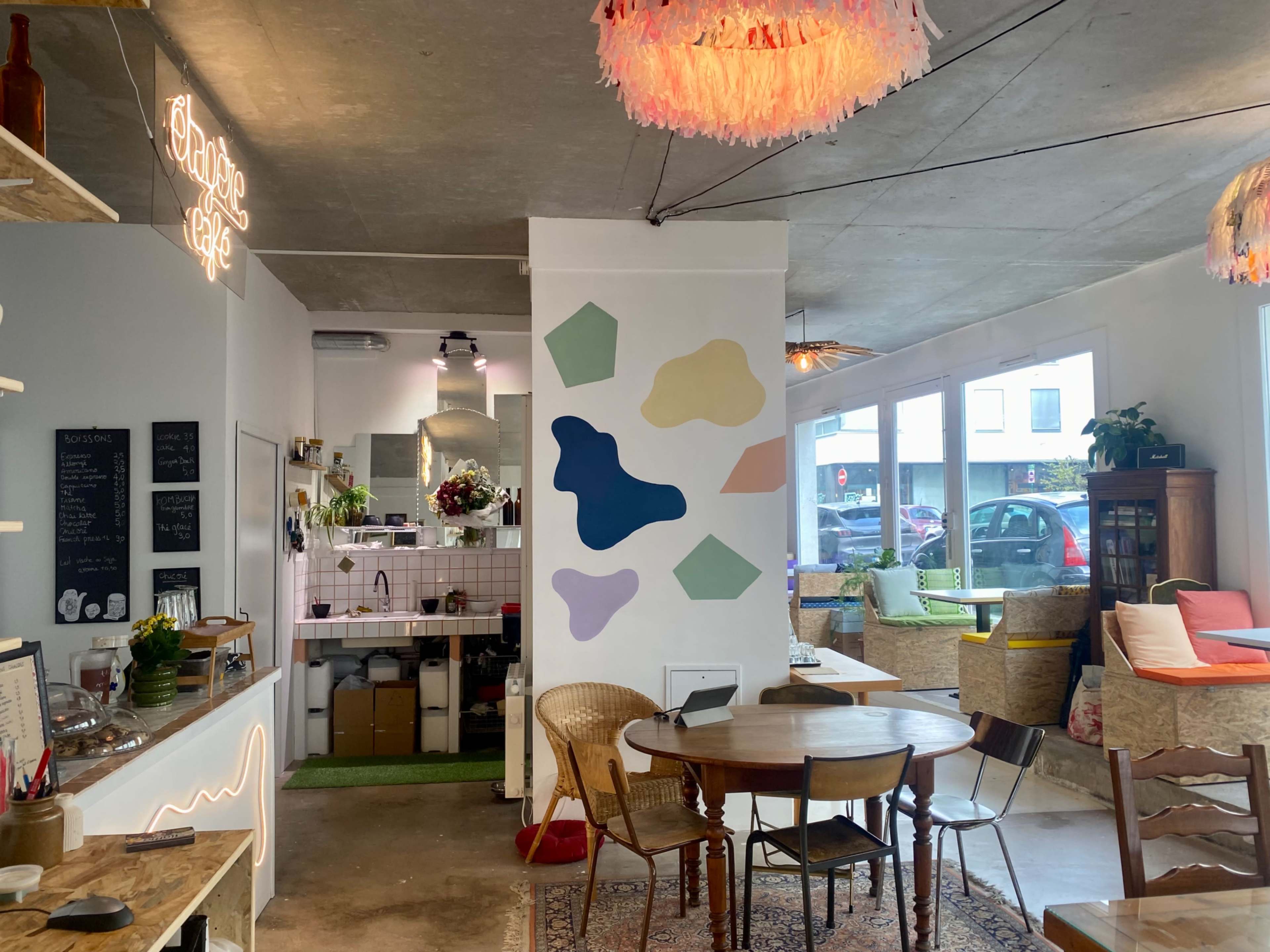 The interior of a café features colorful wall art, a round wooden table, woven chairs, and bright lighting, with a small kitchen visible in the background.