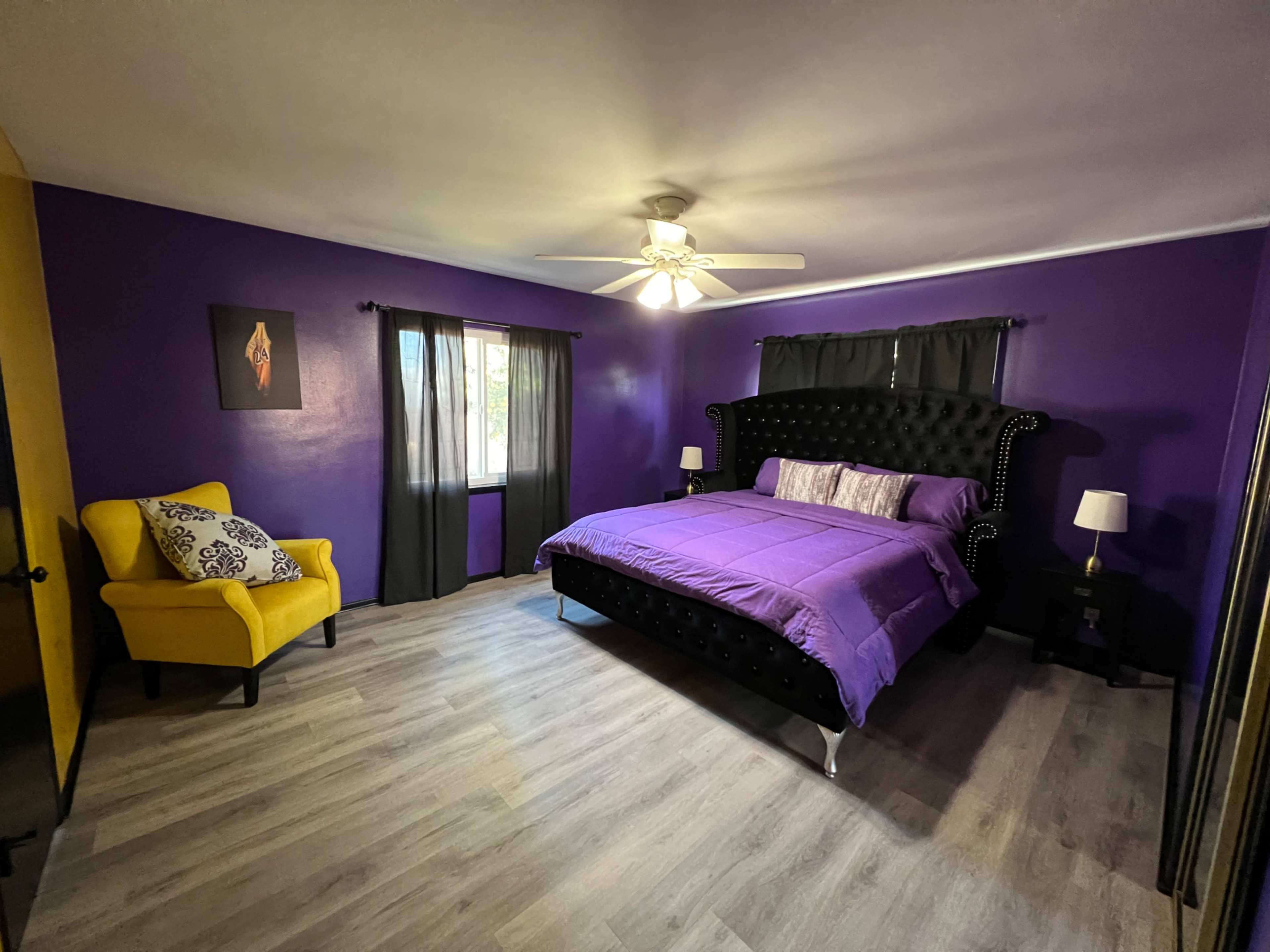 The room features a large bed with a tufted black headboard, purple bedding, a yellow accent chair, and a ceiling fan, all set against purple walls and light-colored flooring.