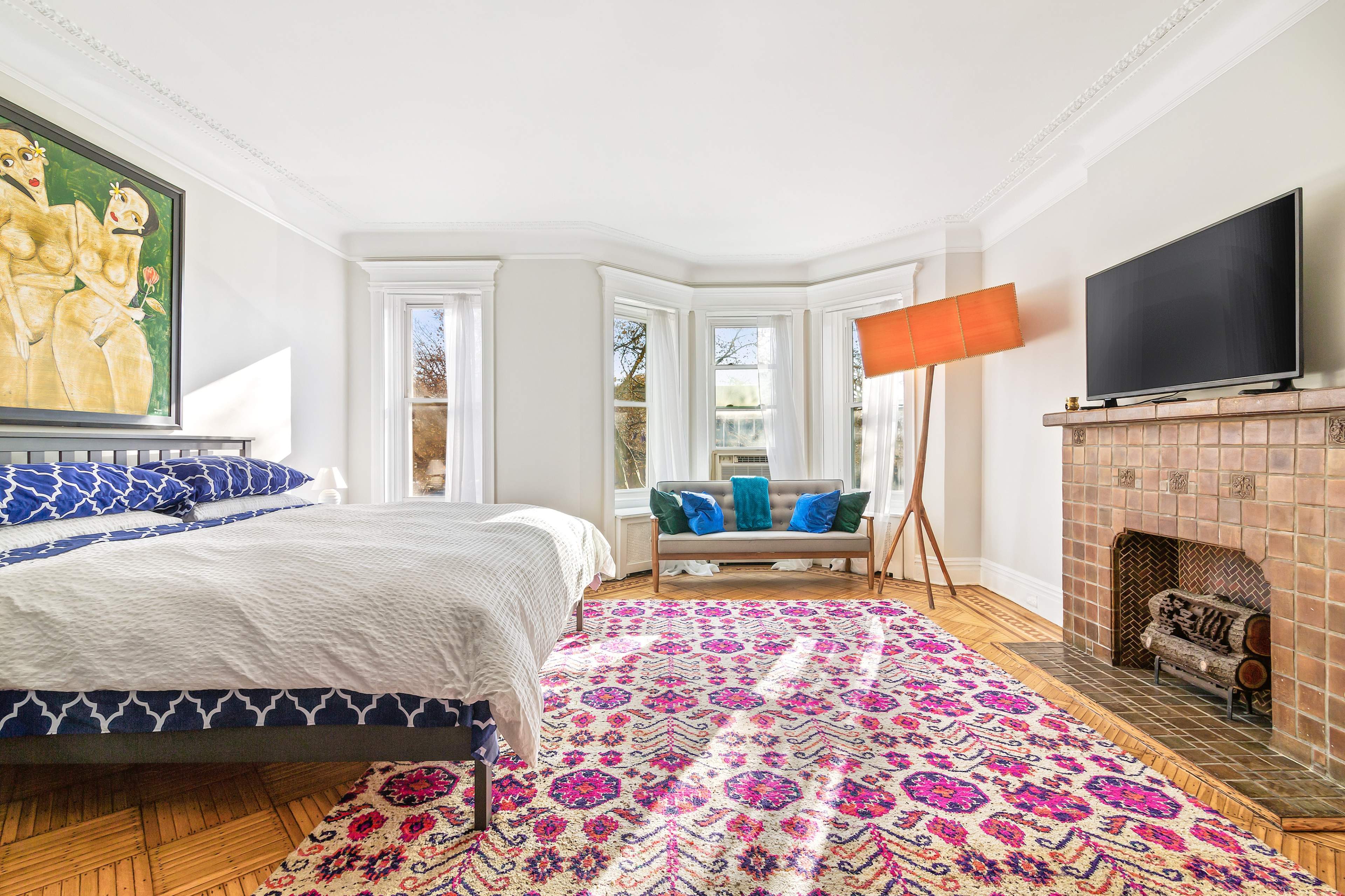 The bedroom features a bed with a patterned blanket, a decorative rug, a modern lamp, and a television mounted above a tiled fireplace, with large windows allowing natural light to fill the space.