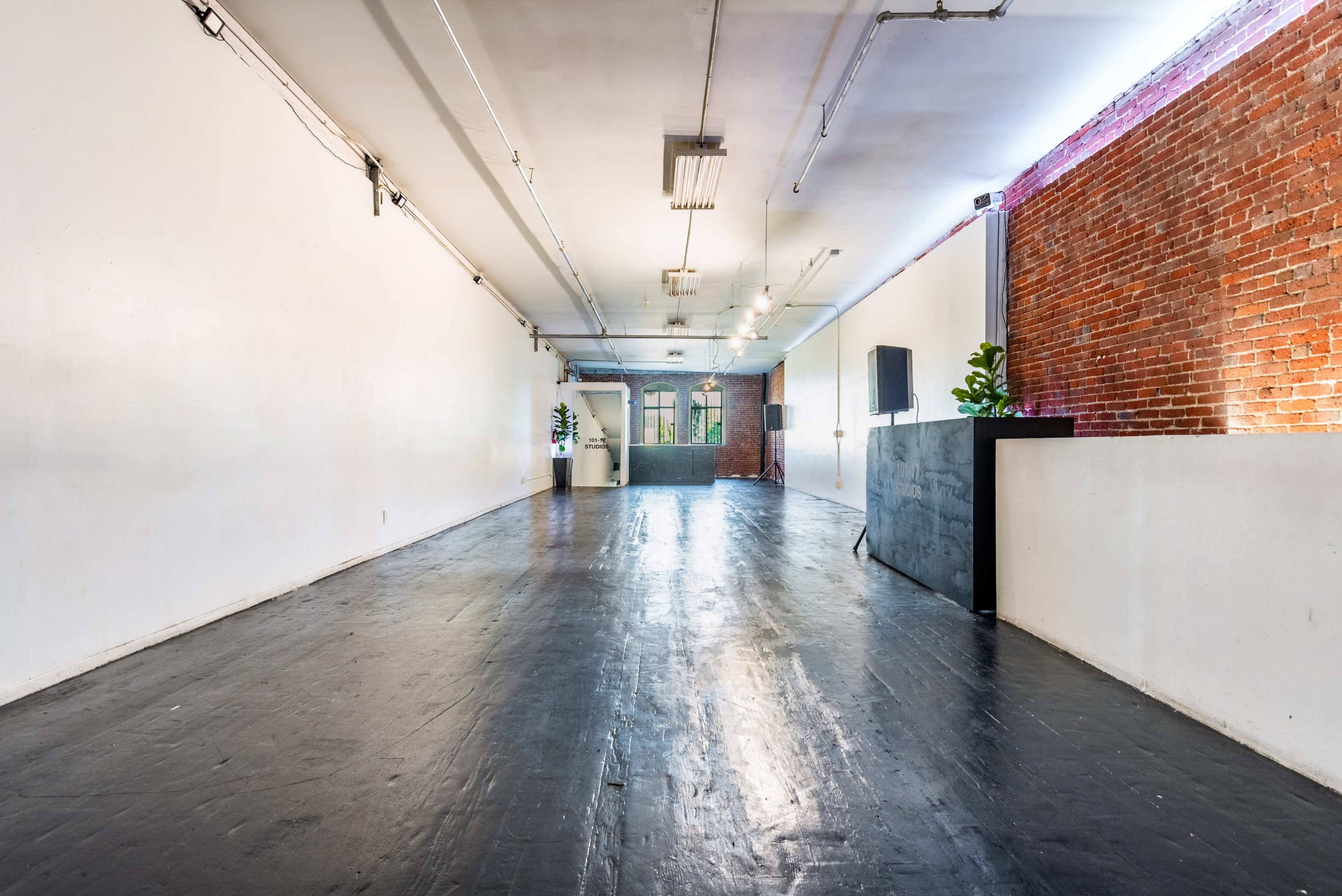 A spacious, empty room with white walls, a dark wooden floor, brick accents, and a small stage at one end.
