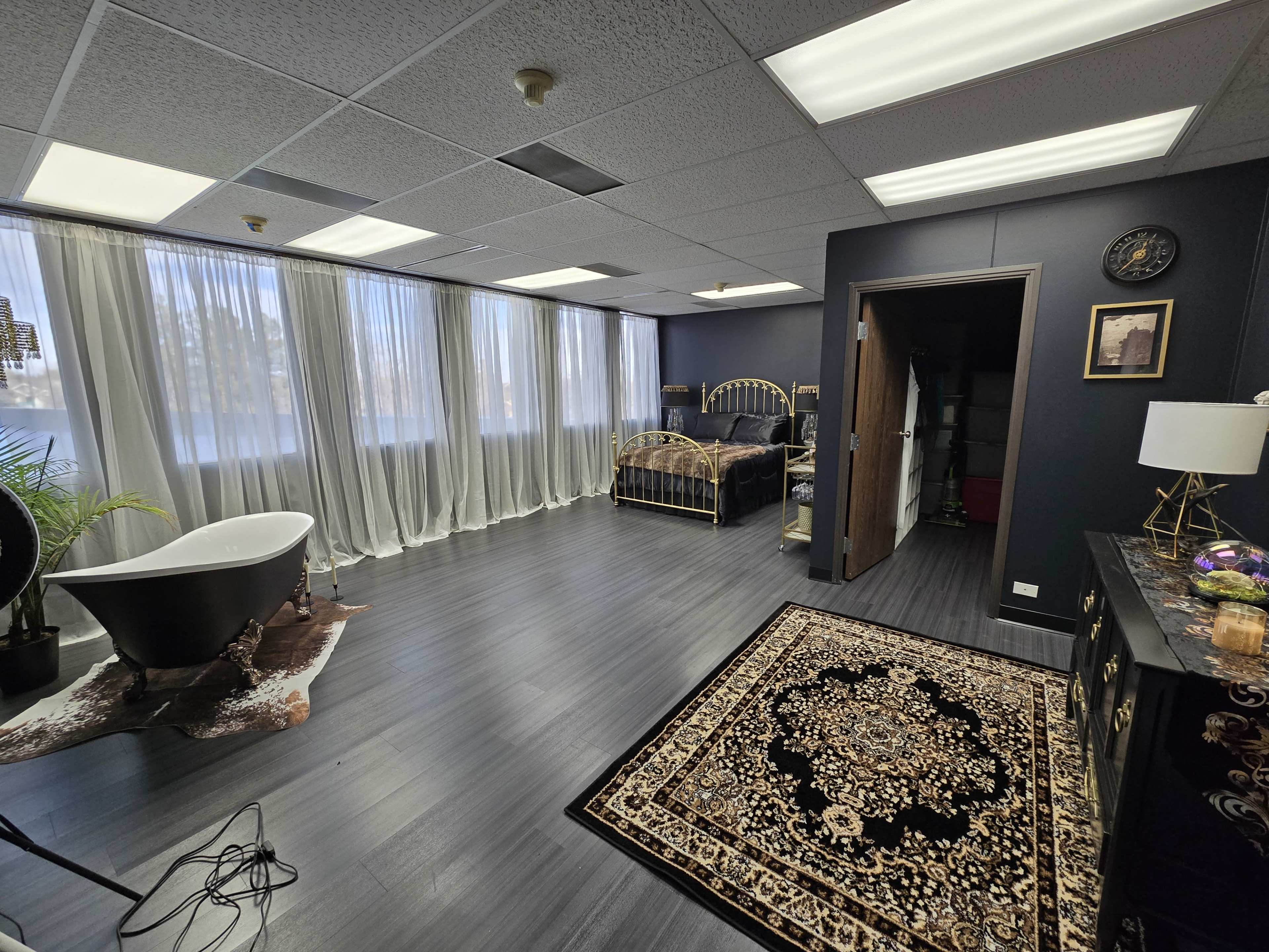 A spacious room with large windows, a bed with a black and gold frame, a black freestanding bathtub, and a decorative rug on the floor.