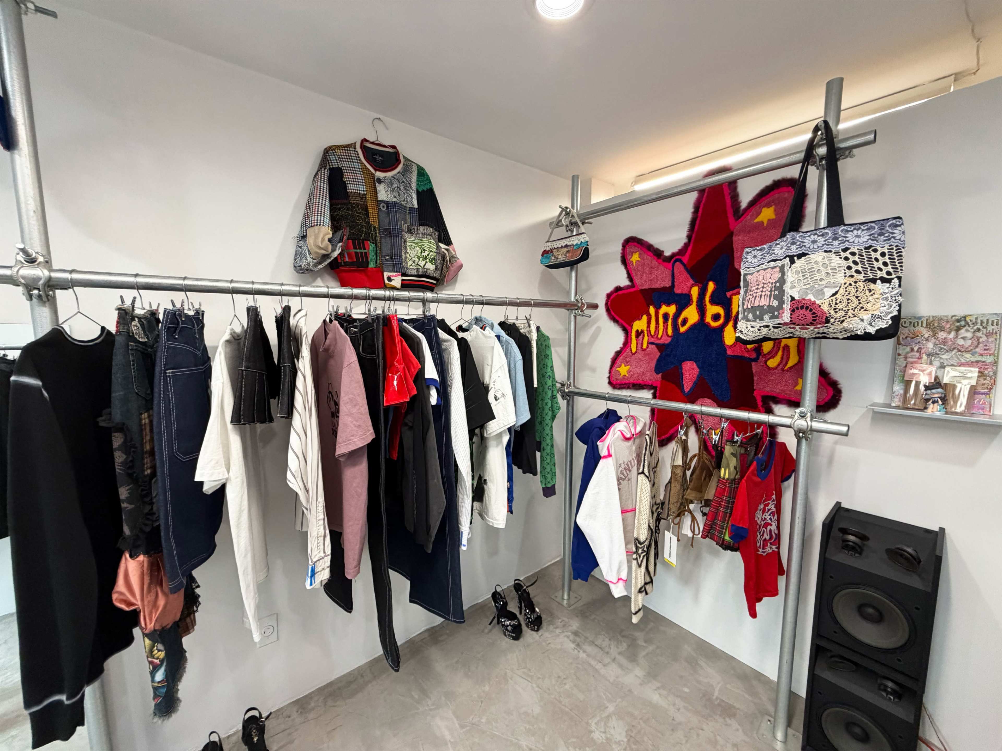 The image shows a clothing display featuring a variety of garments and accessories arranged on metal racks against a white wall.