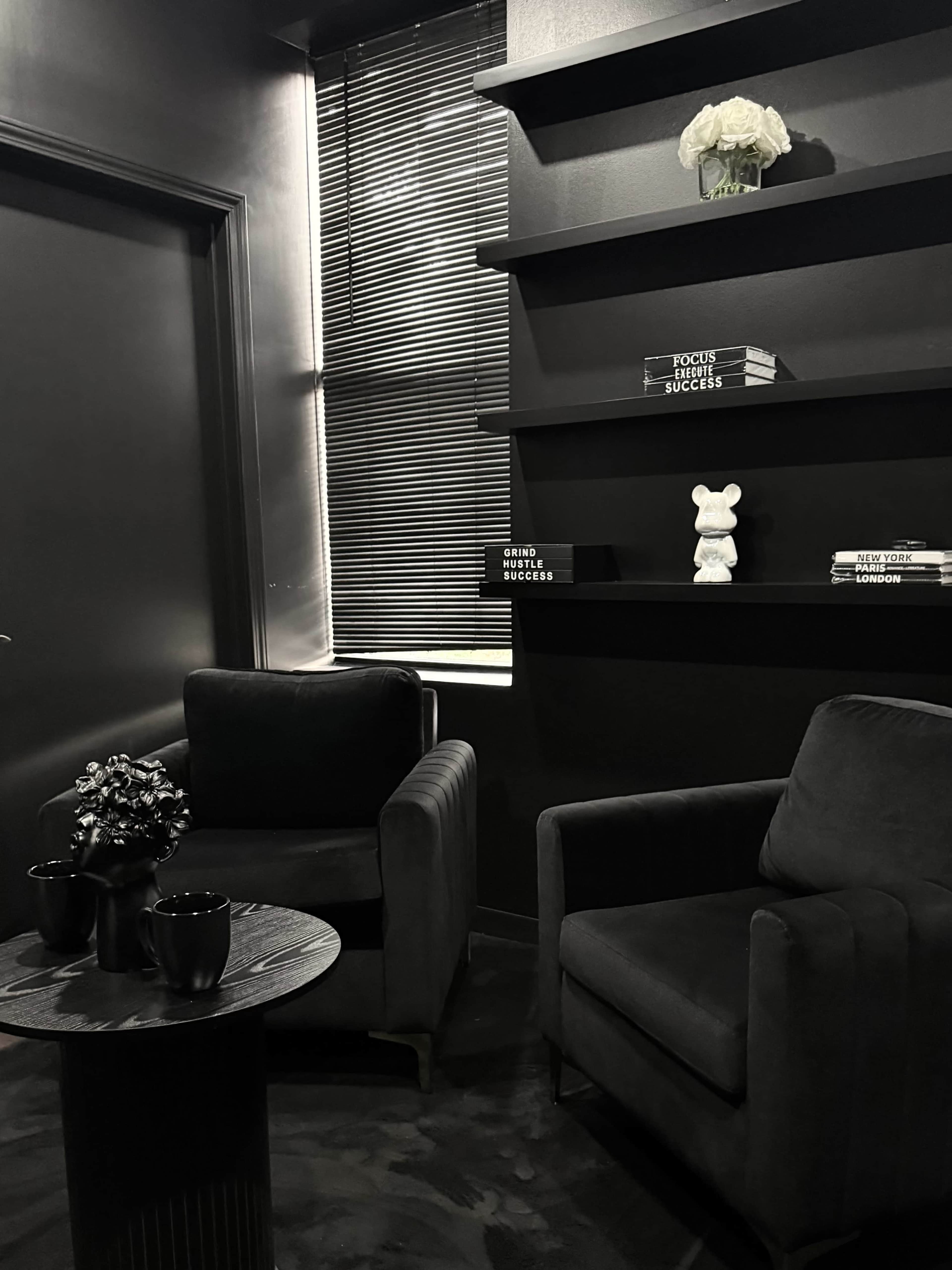 A dimly lit room features two black armchairs facing a circular table, with a shelf displaying books and a decorative item.